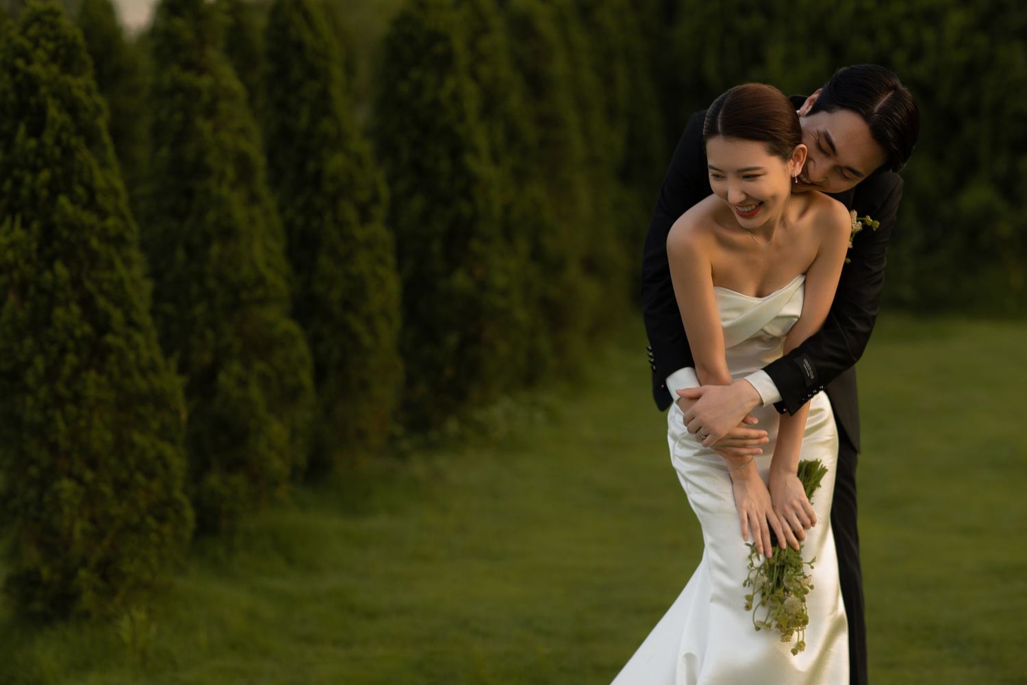 A joyful bride in a white gown laughs as her groom embraces her from behind near tall green cypress trees.