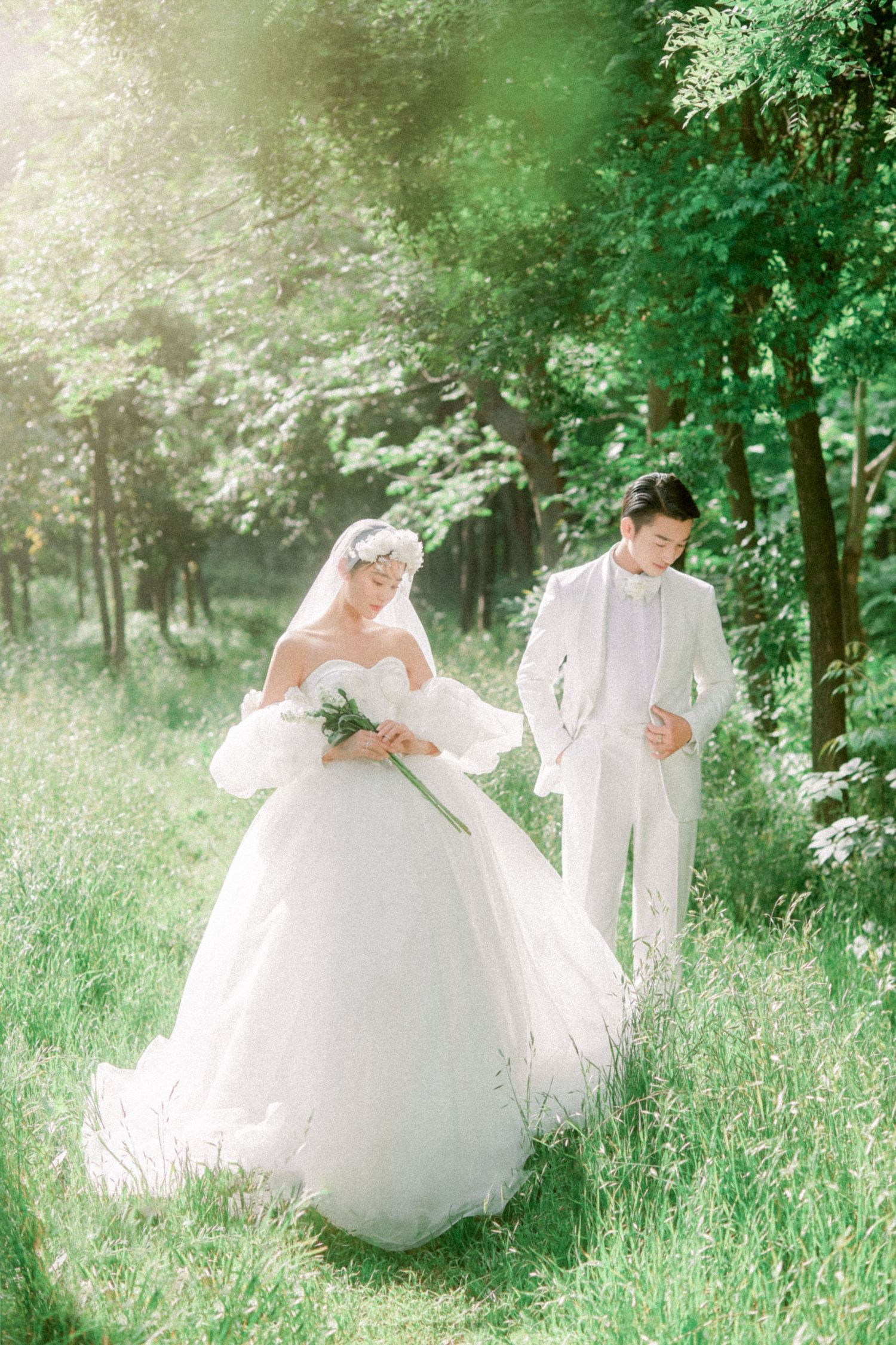 Bride in white ball gown and groom in white suit walk through a sunlit green forest clearing.