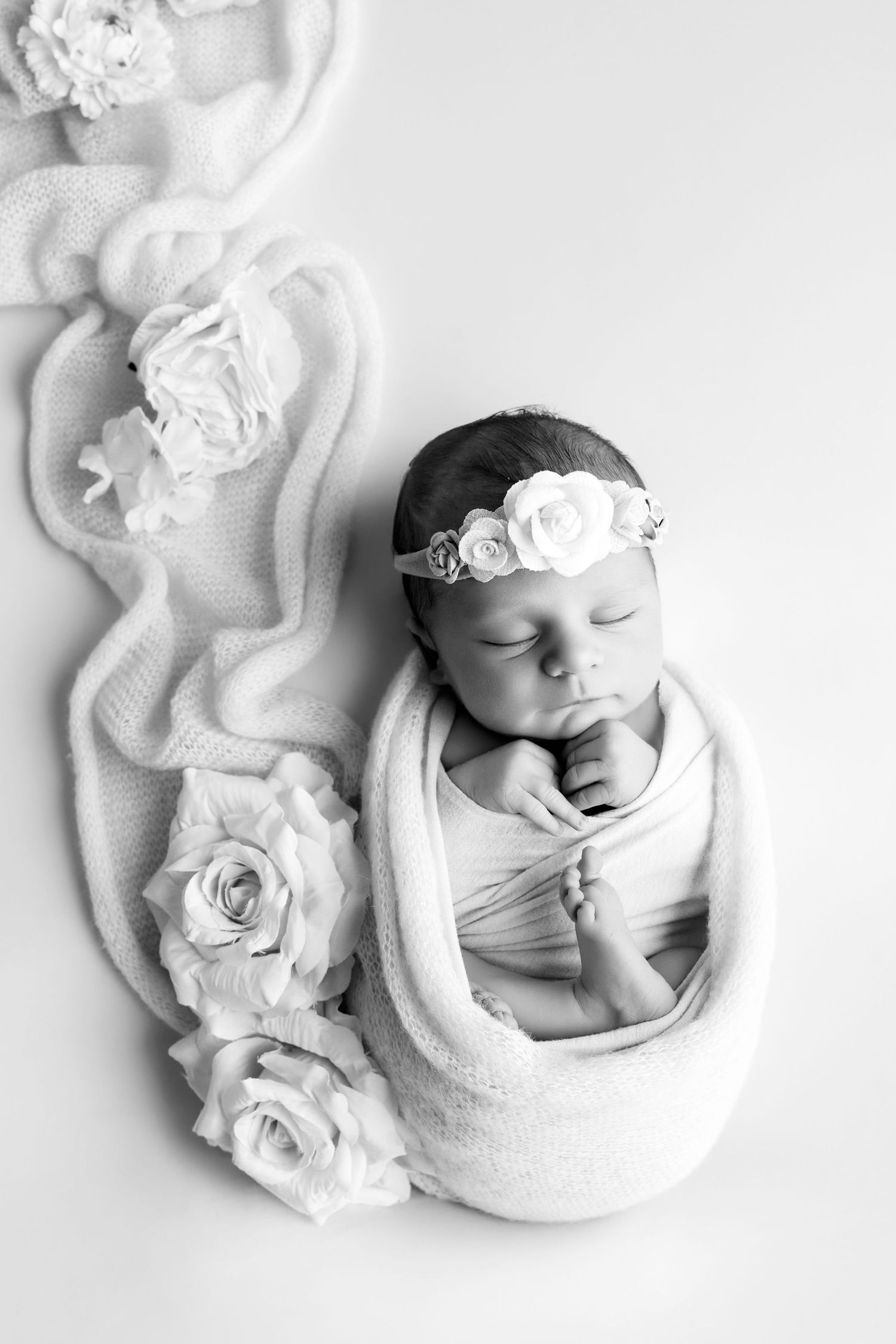 Black and white newborn baby girl swaddled in wrap with floral headband, sleeping peacefully surrounded by roses.