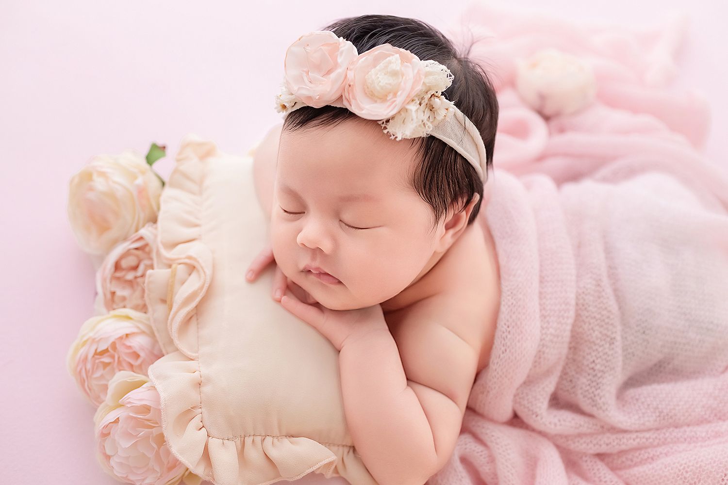 Sleeping newborn baby girl resting on ruffled pillow with pink floral headband and roses.