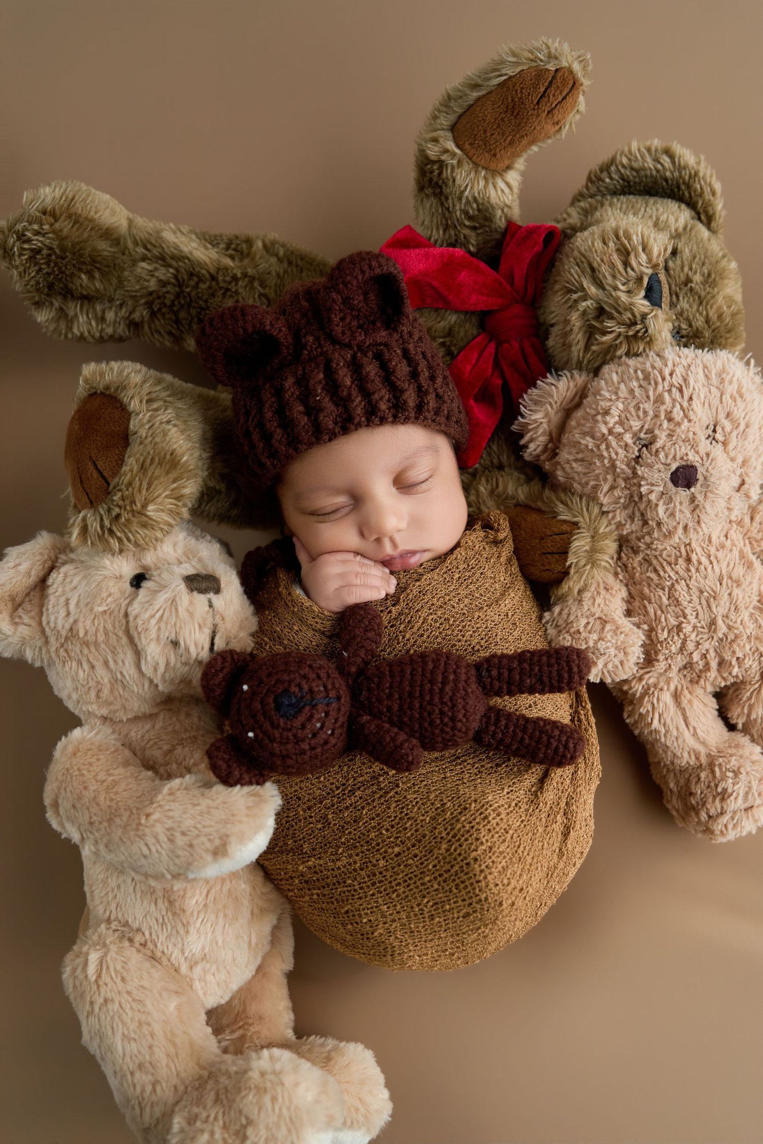Sleeping newborn baby in brown knit hat with red bow, swaddled in burlap, surrounded by stuffed animal toys.