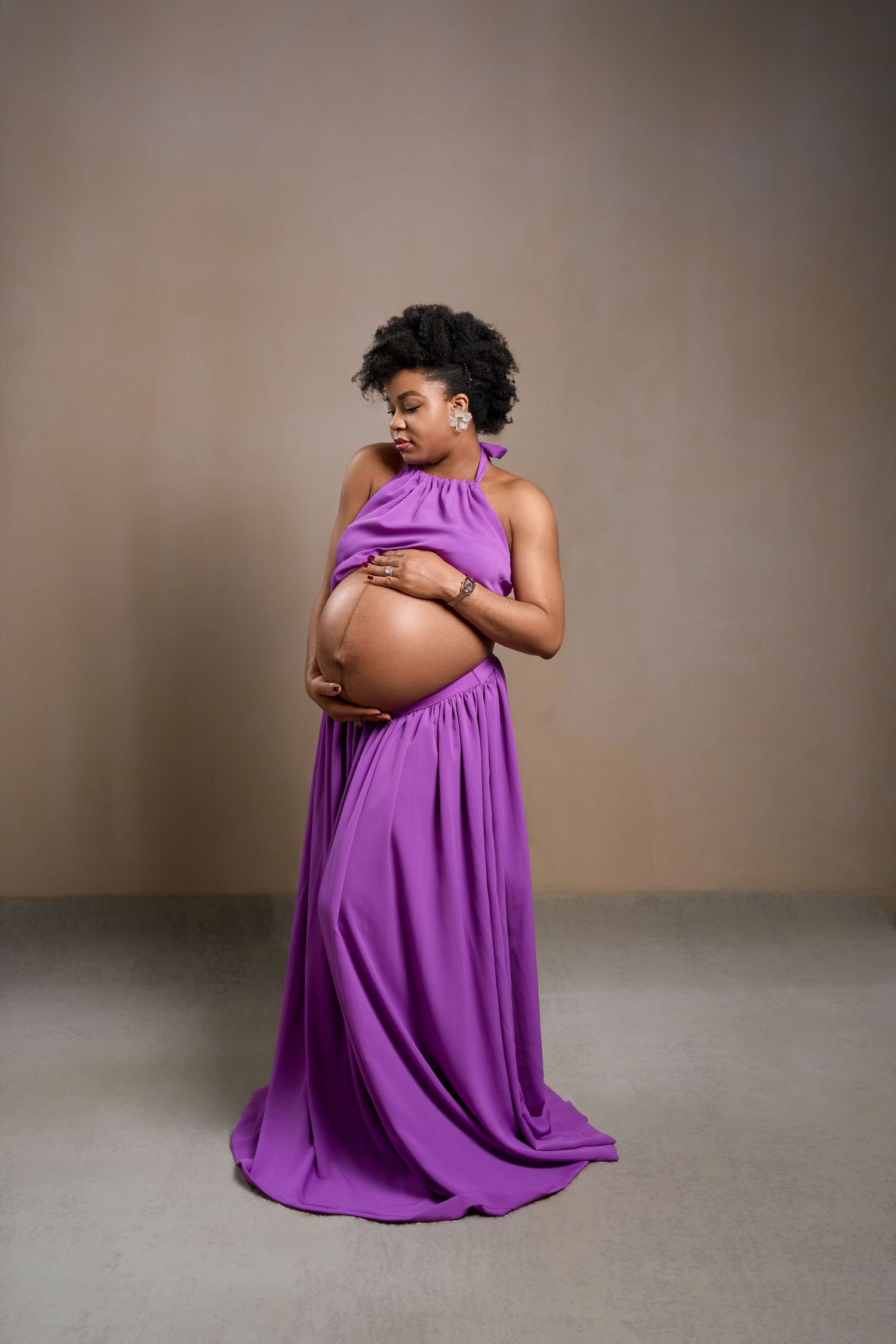 Pregnant woman in purple halter maternity gown cradling baby bump during elegant studio photo session.