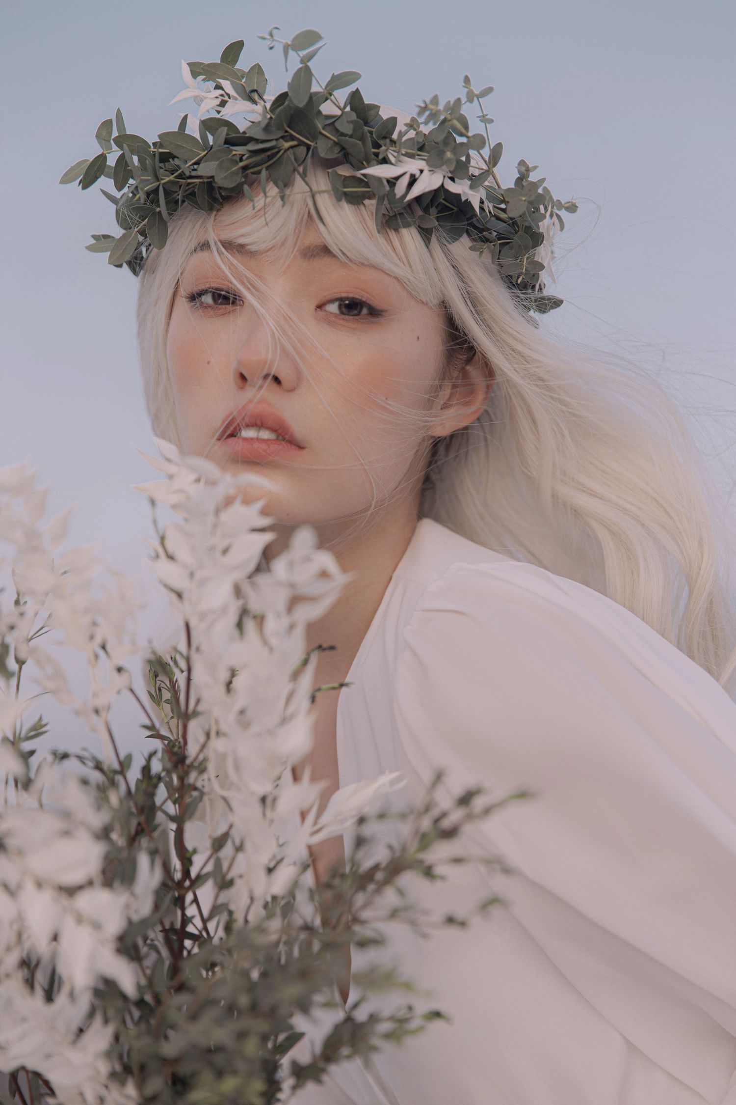 Young woman with platinum blonde hair wearing green leaf crown poses with white flowers in soft light.