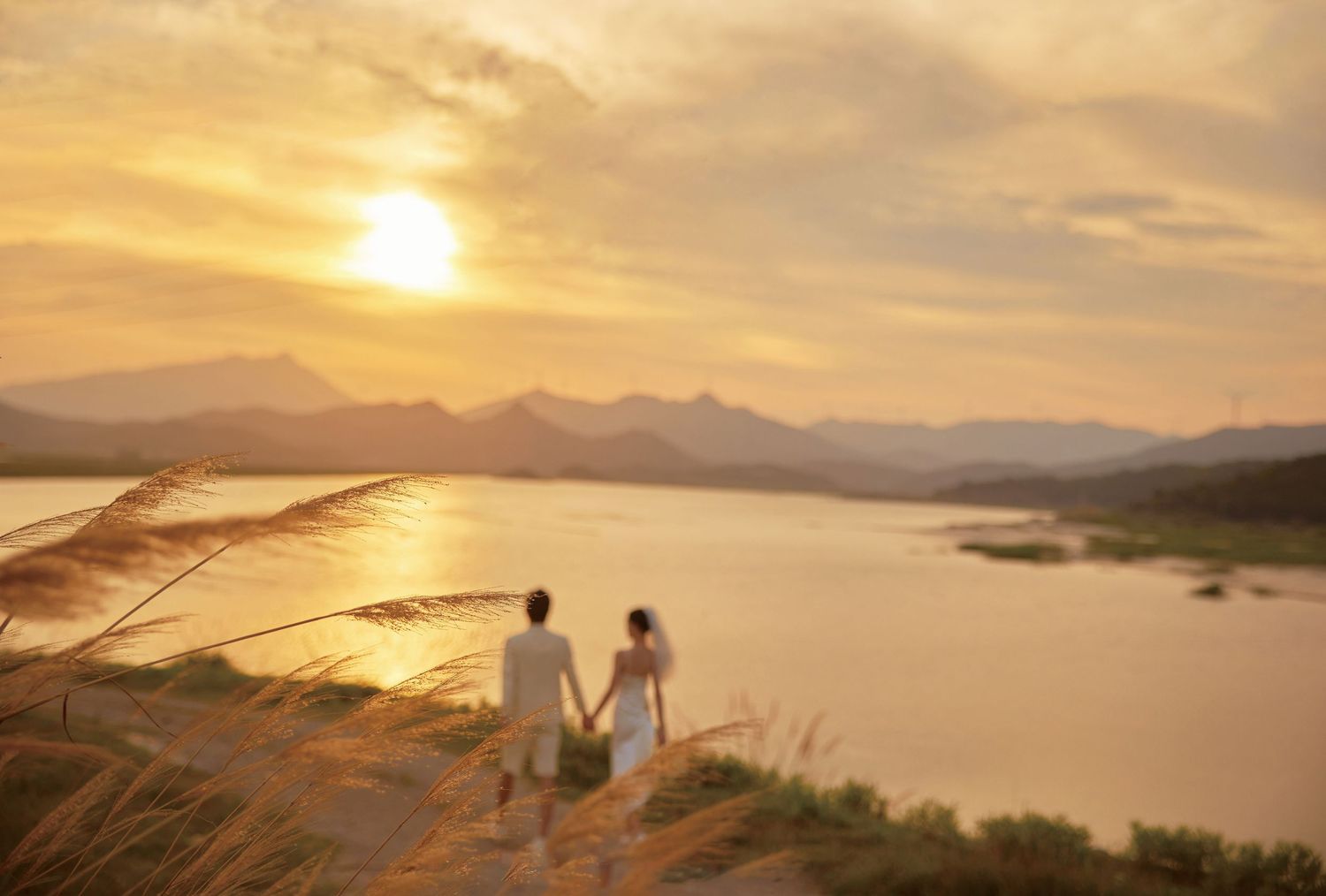 Newlyweds holding hands overlooking a serene river landscape during a breathtaking golden sunset.