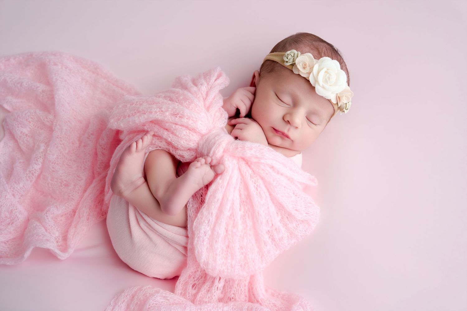 Sleeping newborn baby girl wrapped in pink lace fabric with floral headband on pink background.
