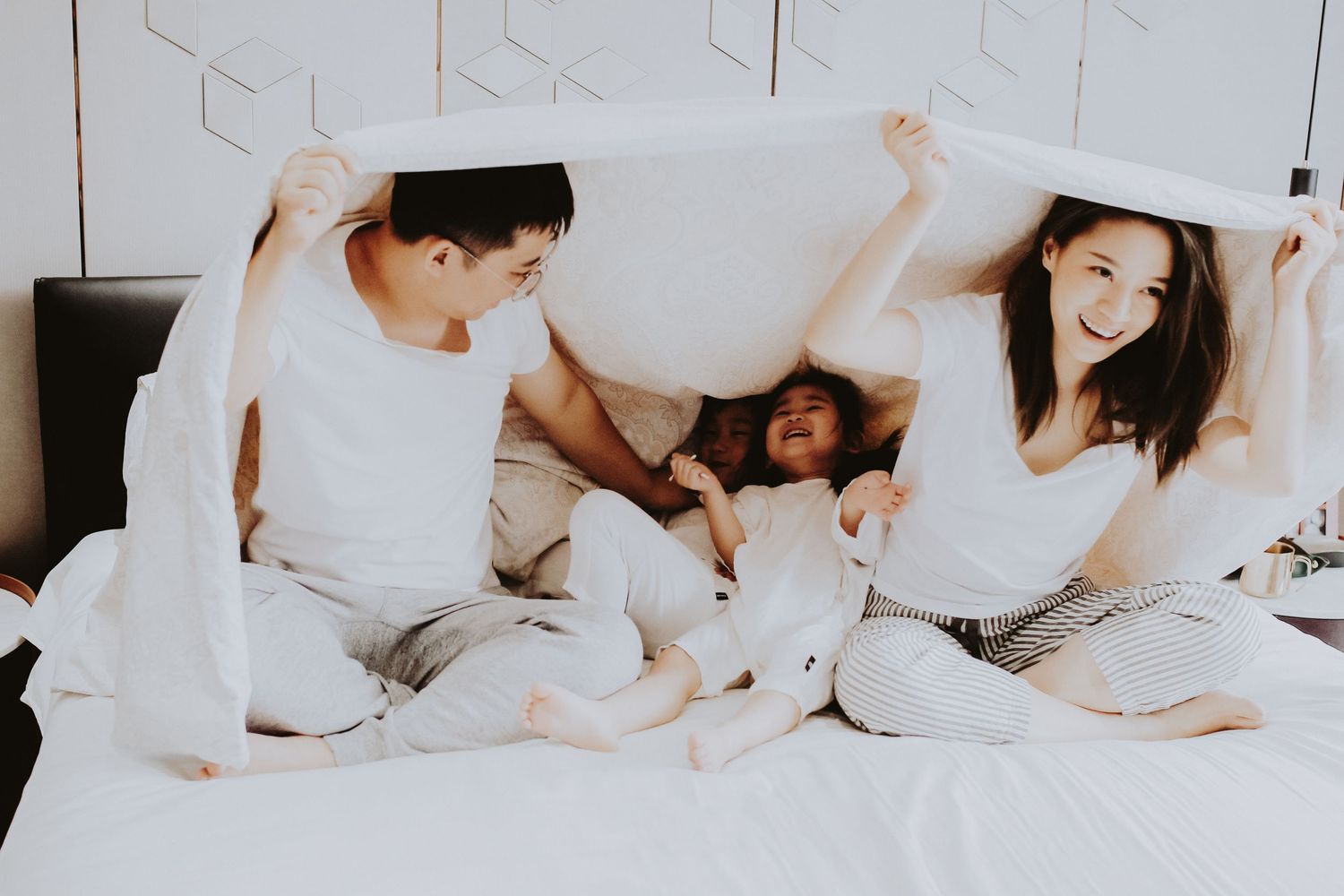 A joyful Asian family of four plays under a white blanket on a bed, laughing and having fun together.