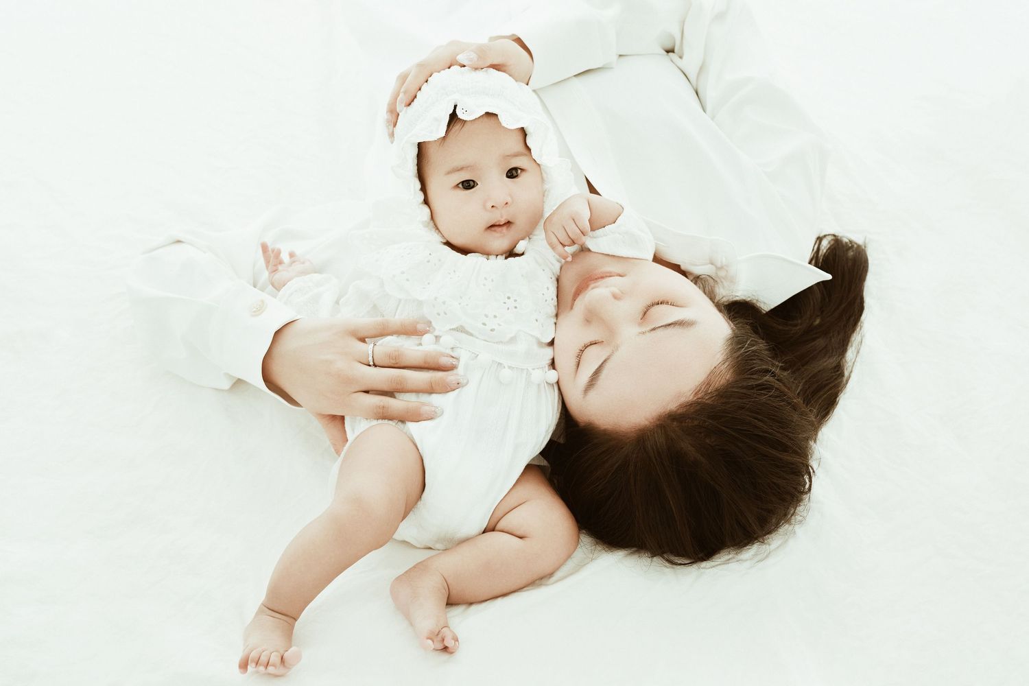 Asian mother lying on white surface smiling at her baby girl in white dress and bow headband.