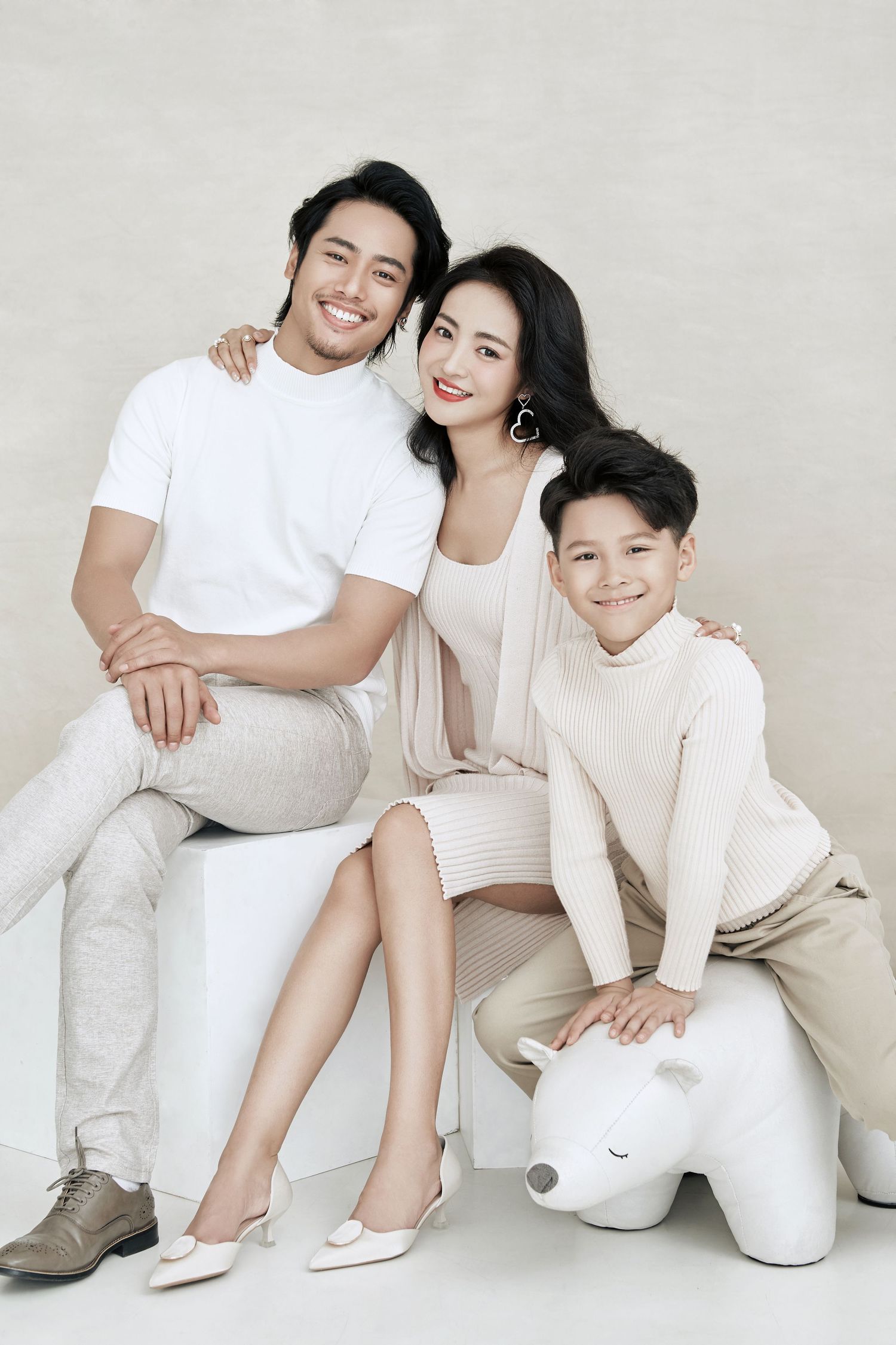 A smiling Asian family of three posing together in coordinated neutral outfits for a studio portrait.