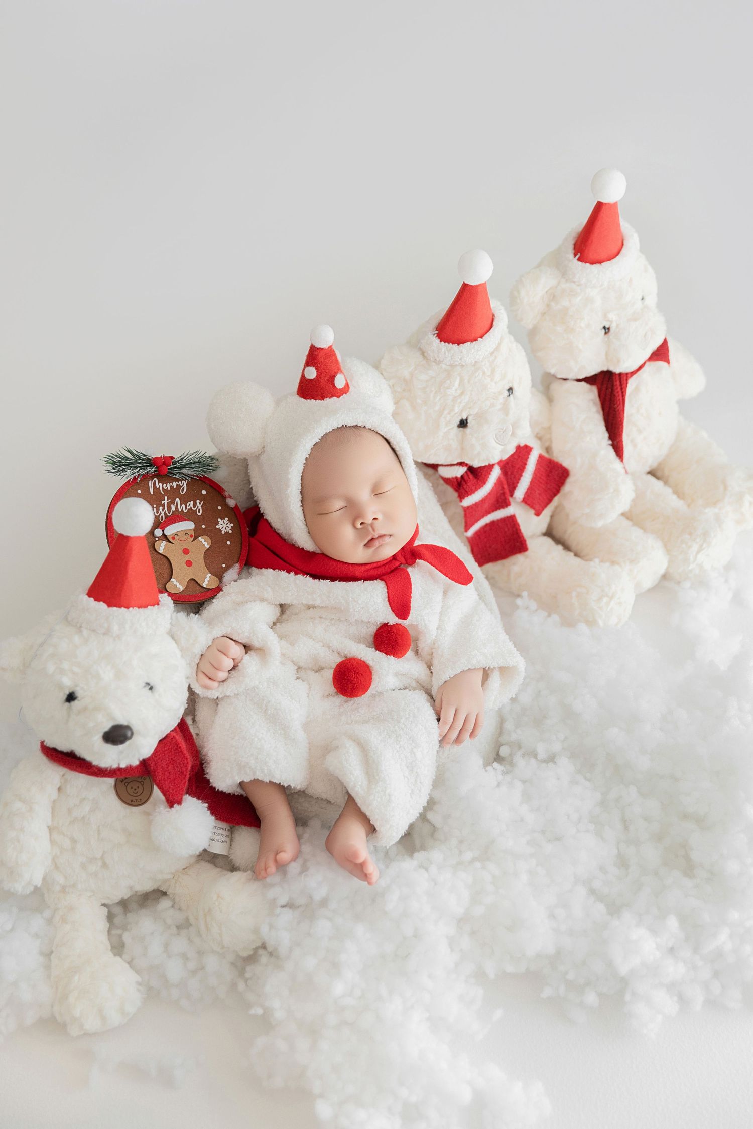 Peaceful newborn in Santa outfit resting among white polar bear plush toys with red hats on snowy surface.