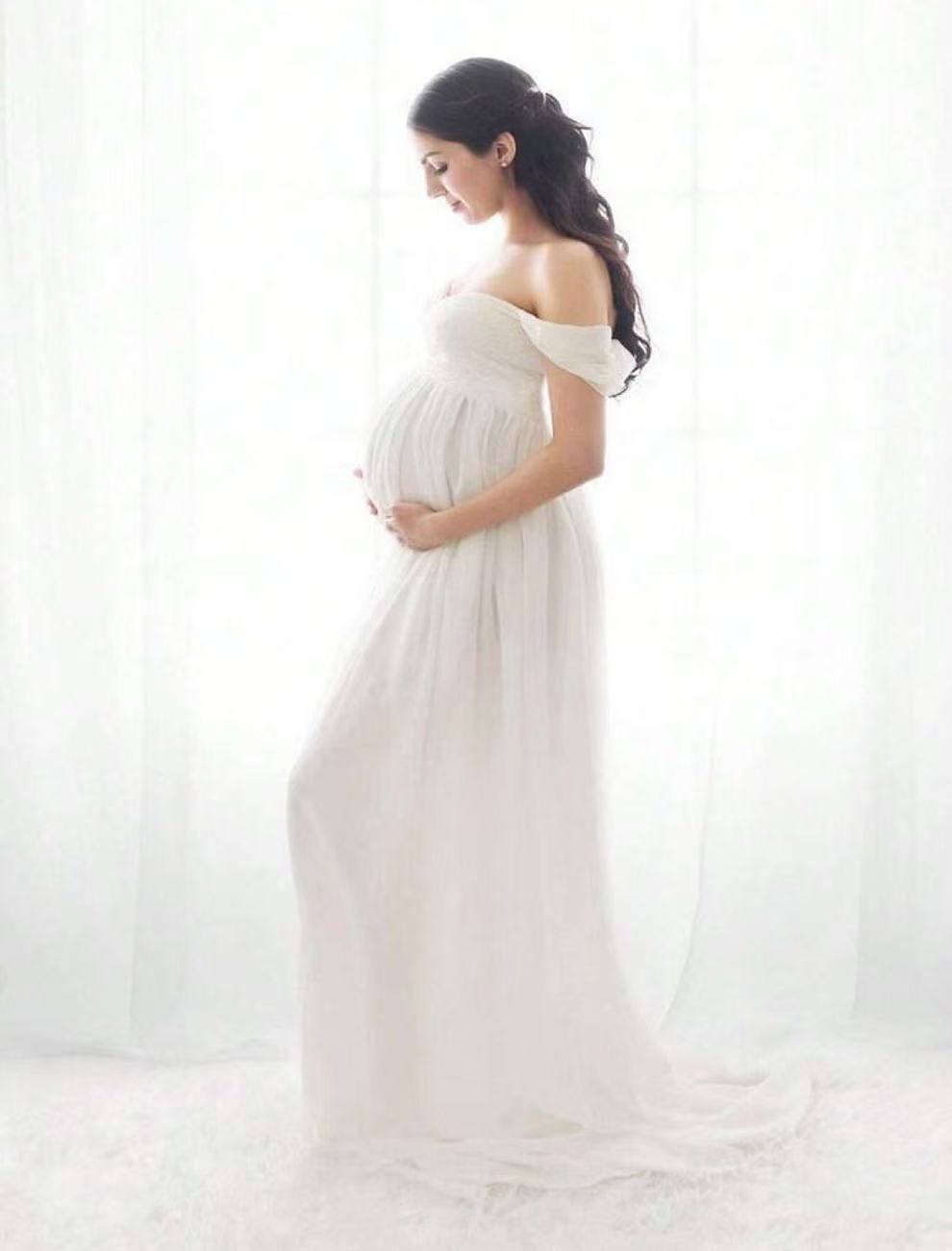 Pregnant woman in elegant white off-shoulder chiffon maternity gown posing softly in front of sheer curtains.