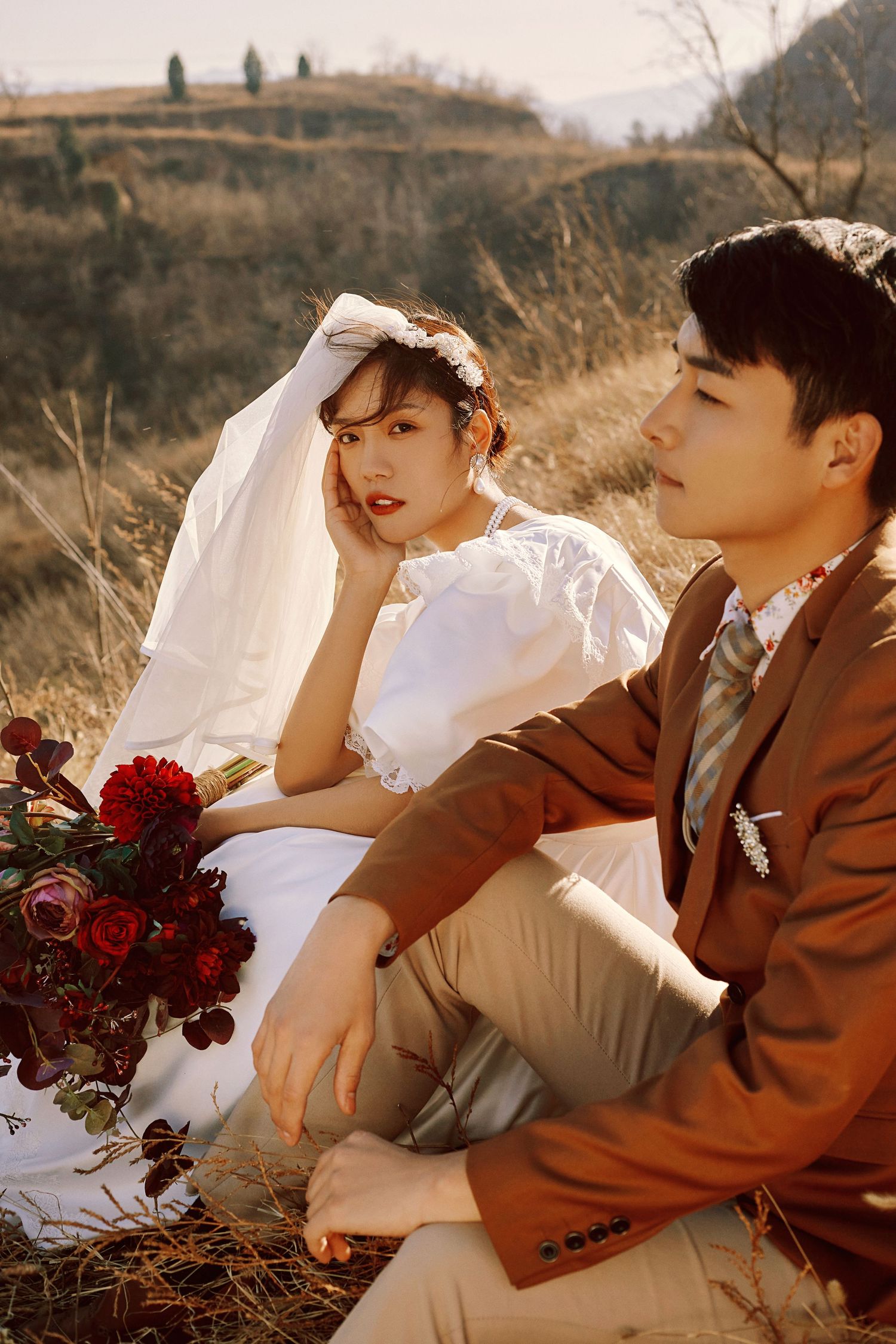 Asian couple in wedding attire sit together in dry grass field holding red roses at golden hour.