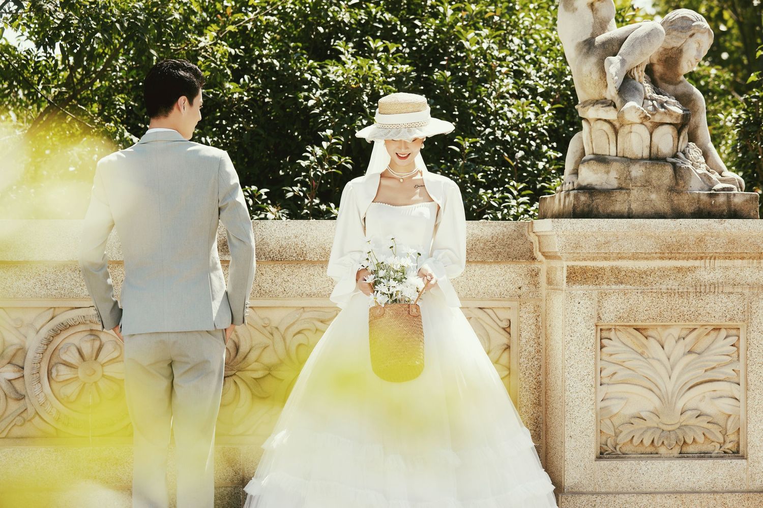 Bride in white ballgown and straw hat stands at stone terrace as groom walks nearby in garden setting.