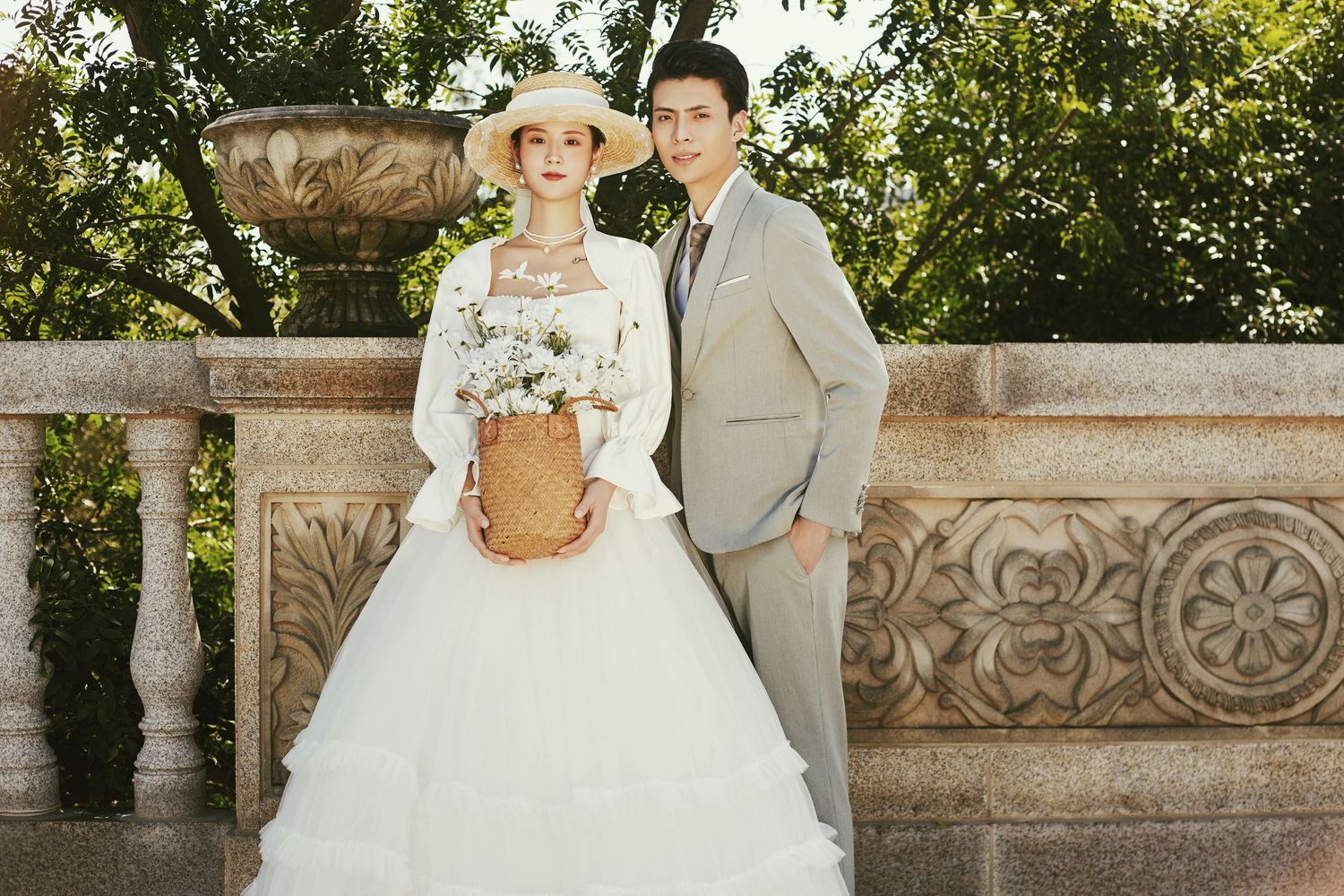 Wedding couple poses together at stone balcony, bride in white dress and hat holding wicker basket bouquet.
