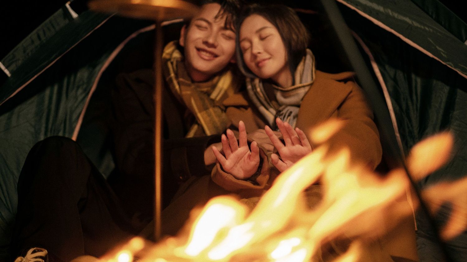 Couple warming hands by campfire inside tent at night.