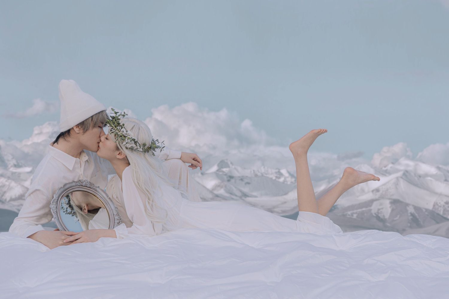 A romantic couple in white attire shares a kiss while lying on a snowy white surface with mountain backdrop.
