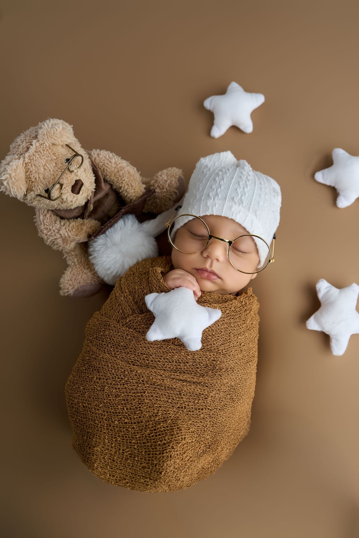 Newborn baby sleeping wrapped in brown knit swaddle with white hat, glasses, teddy bear, and star pillows.