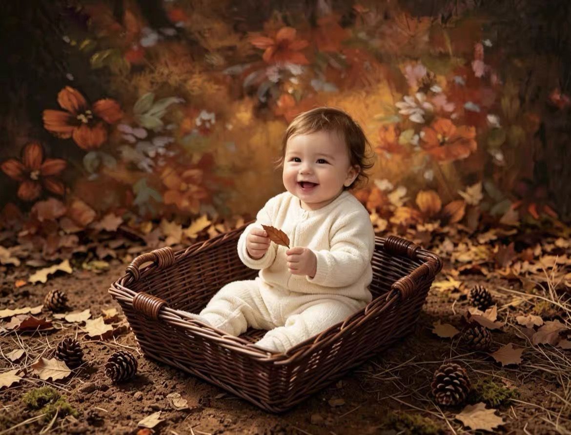 Smiling baby in white outfit sitting in wicker basket surrounded by autumn leaves and pinecones on fall backdrop.