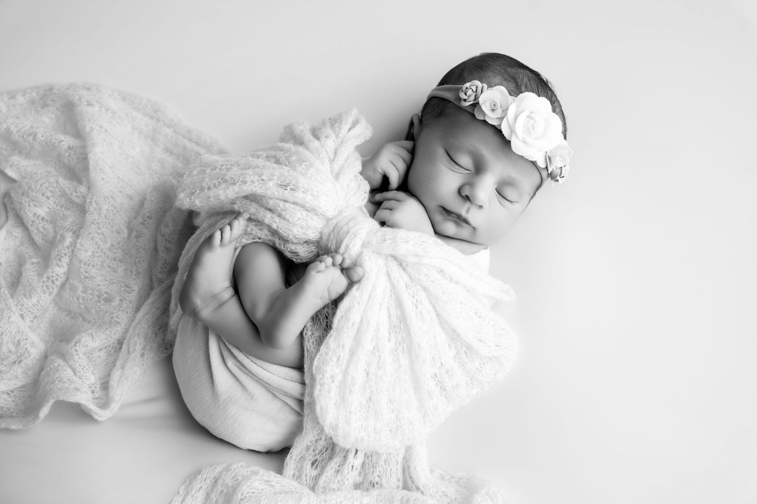 Black and white newborn photo of sleeping baby wrapped in lace, wearing floral headband.