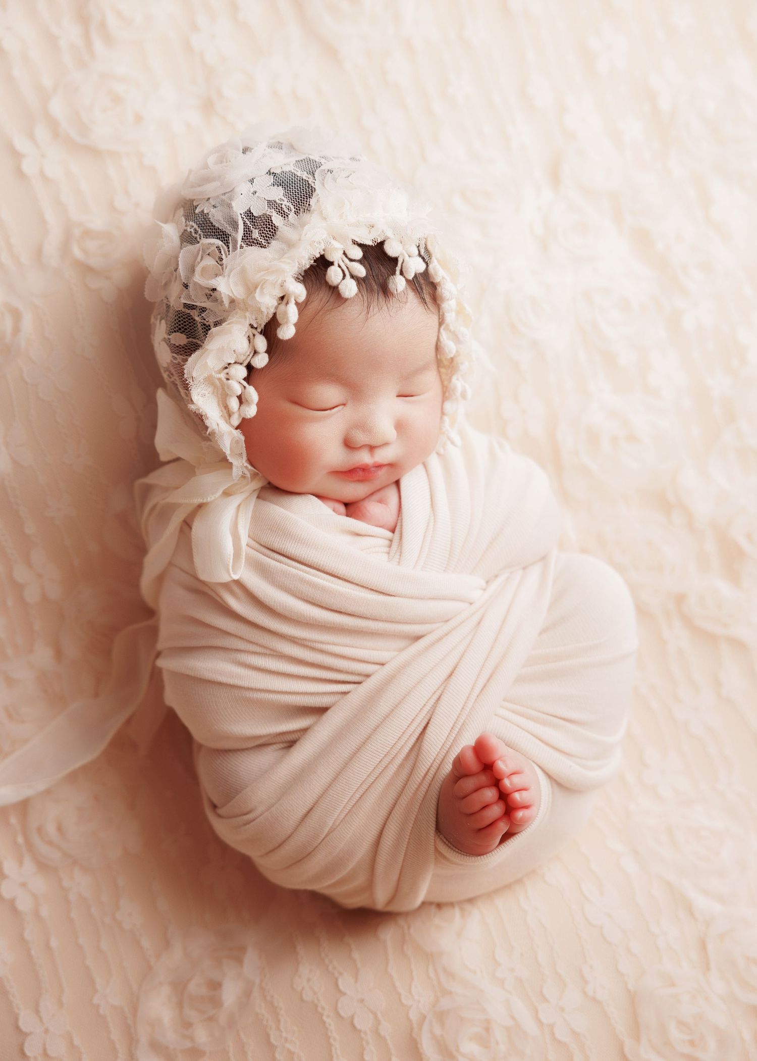 Swaddled newborn baby in cream wrap wearing delicate lace bonnet, smiling.