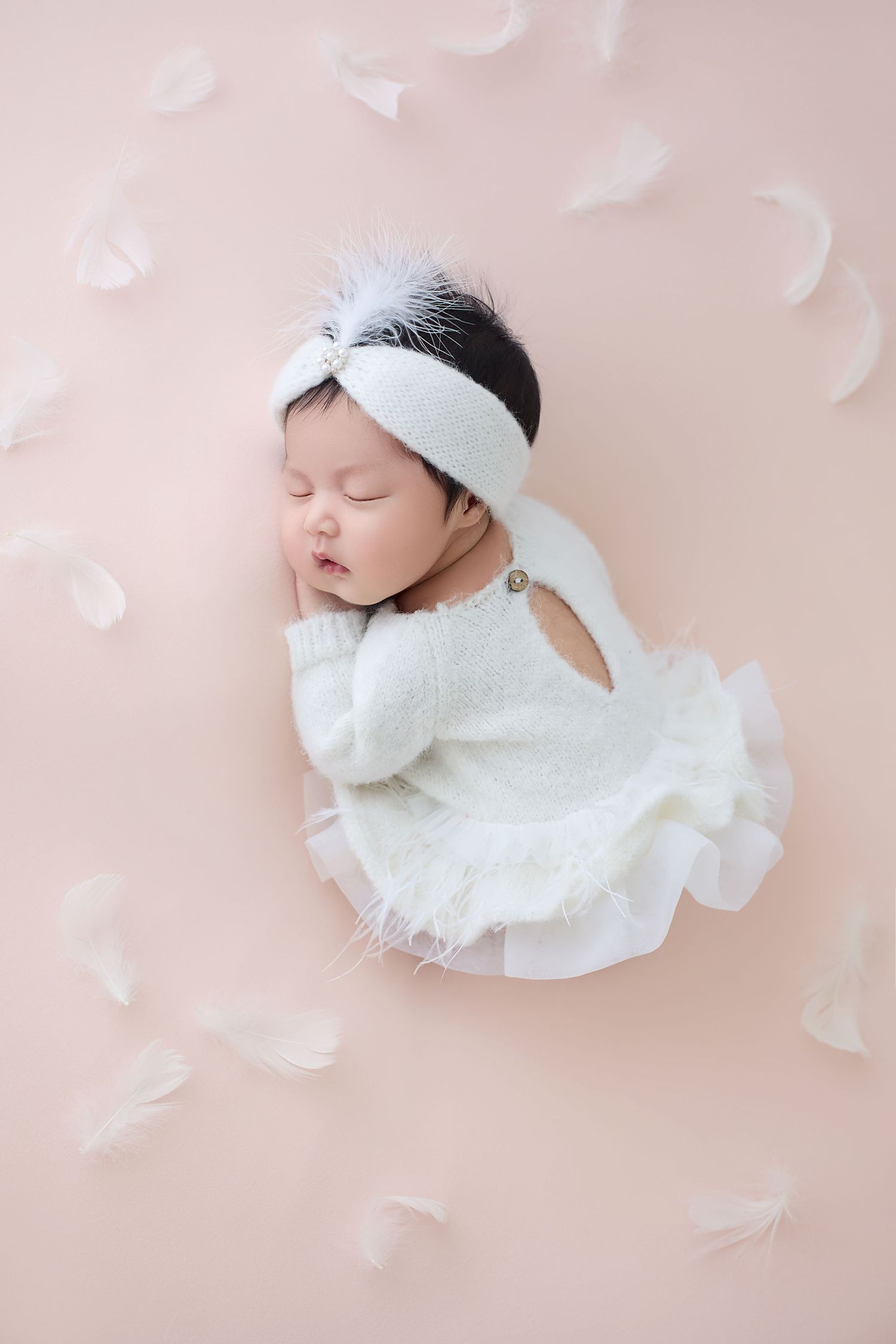 Sleeping newborn baby in white knit outfit with feather headband surrounded by white feathers on pink background.