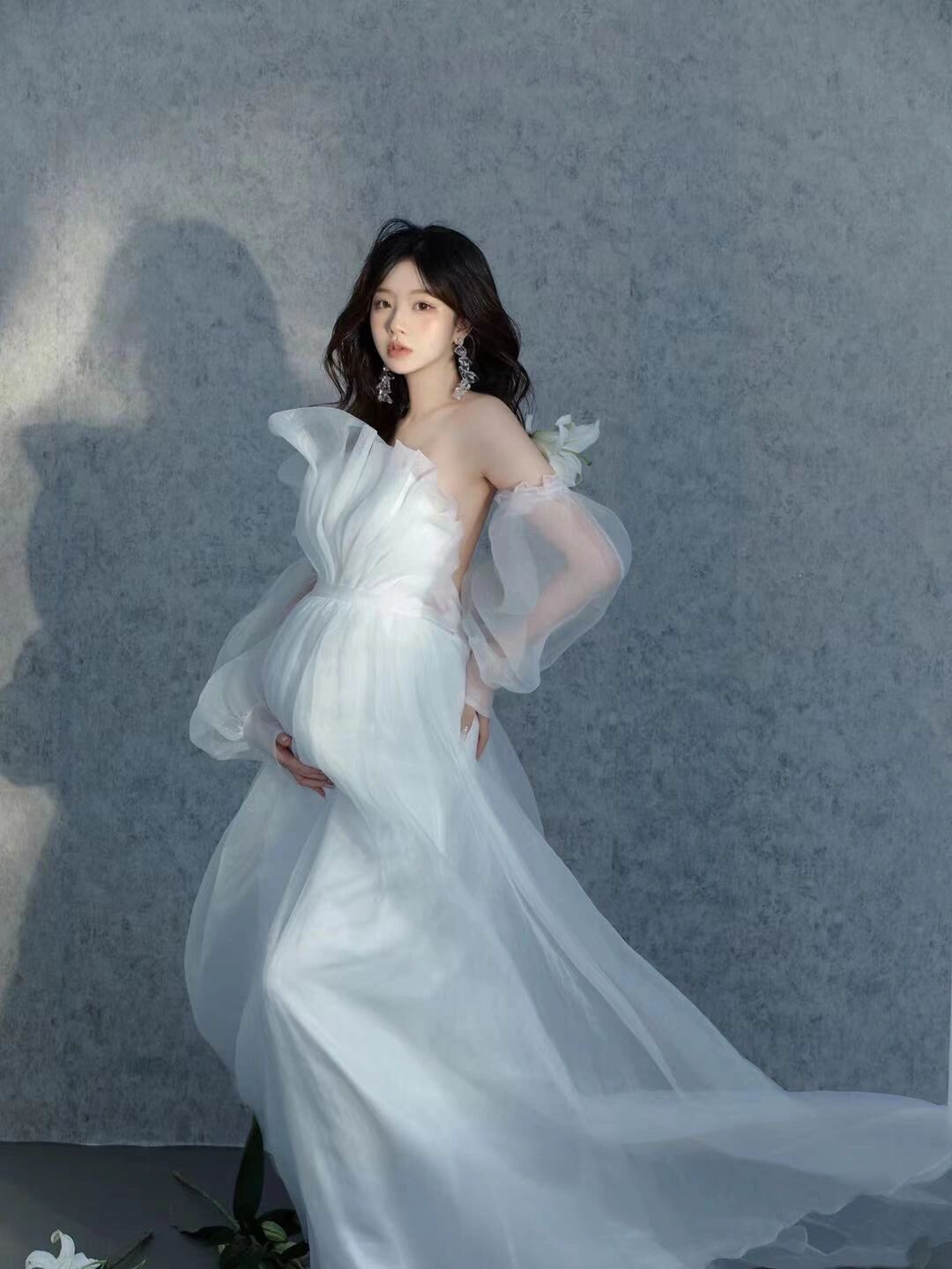 Pregnant woman in elegant white tulle off-shoulder maternity gown posing gracefully with white lily flowers.