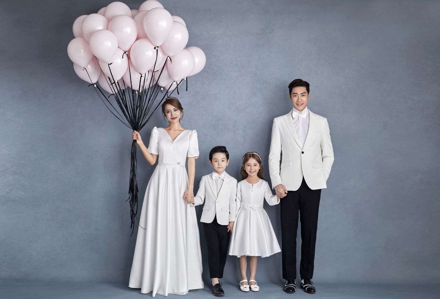 Elegant Asian family of four in formal white outfits pose with pink balloons in a stylish studio portrait.