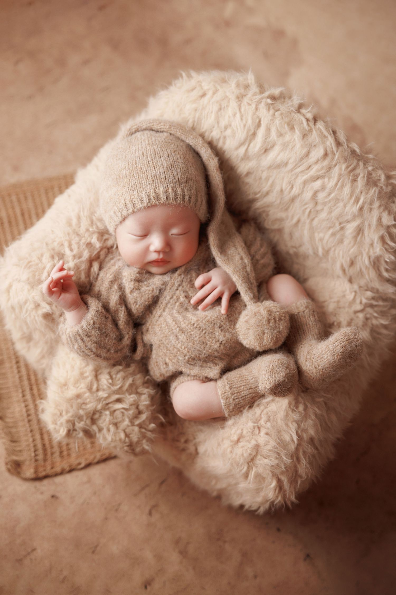 Peaceful newborn baby in tan knit romper and bunny hat resting in cozy cream fluffy bowl on beige background.