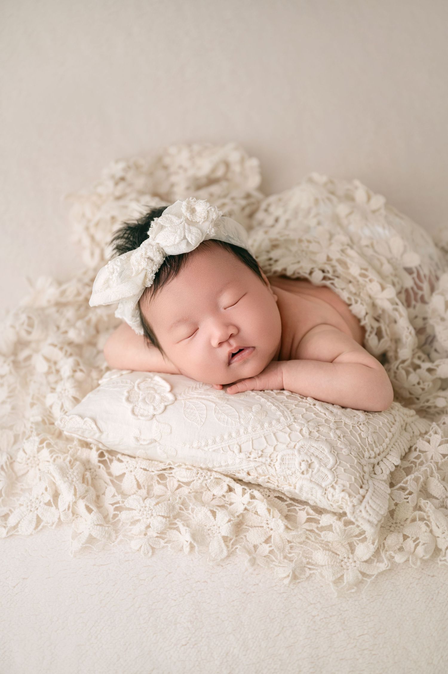 Sleeping newborn baby girl on lace pillow with white bow headband.
