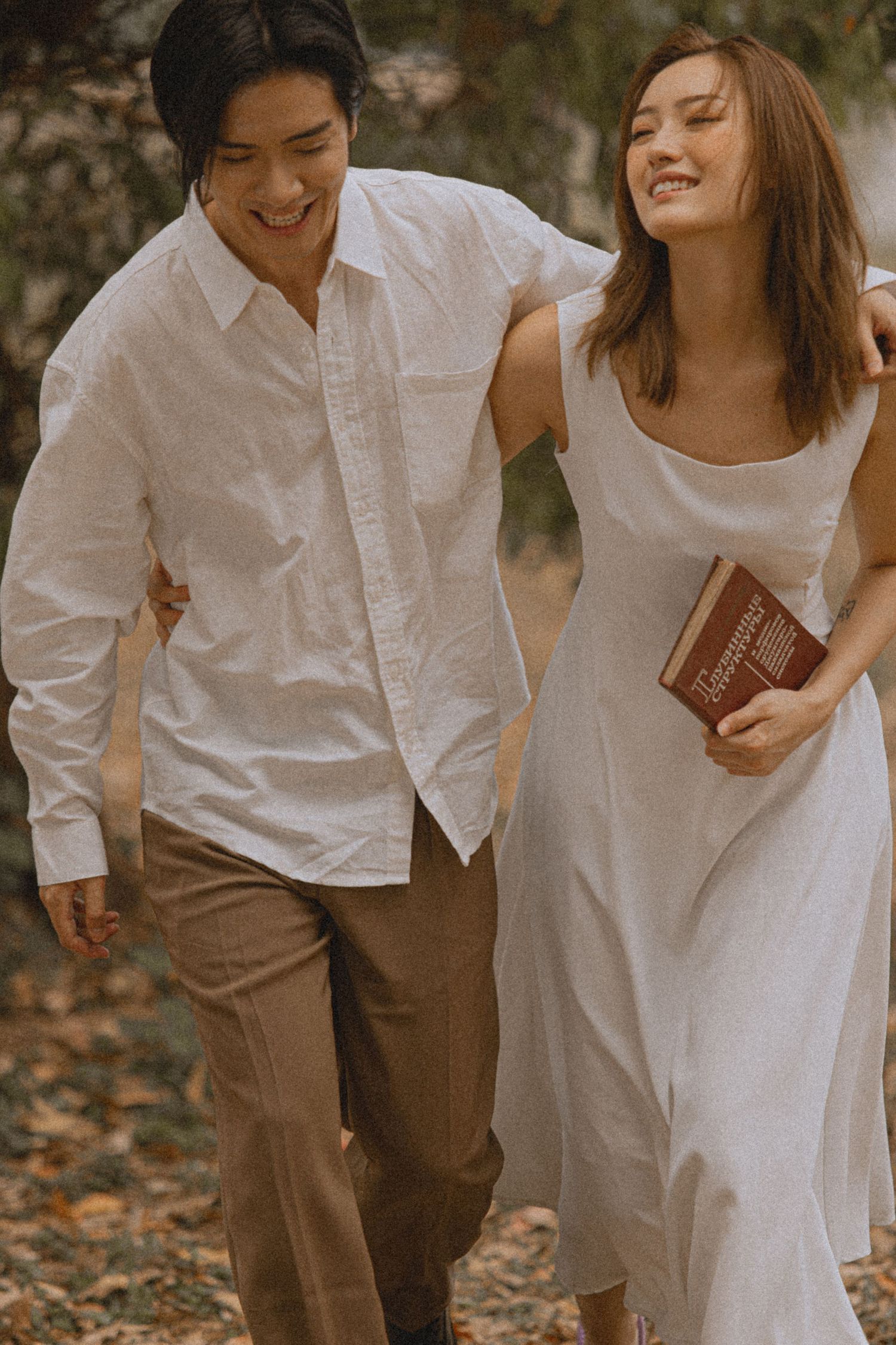 Smiling couple walking together outdoors, woman in white dress holding a book, man in white shirt and tan pants.