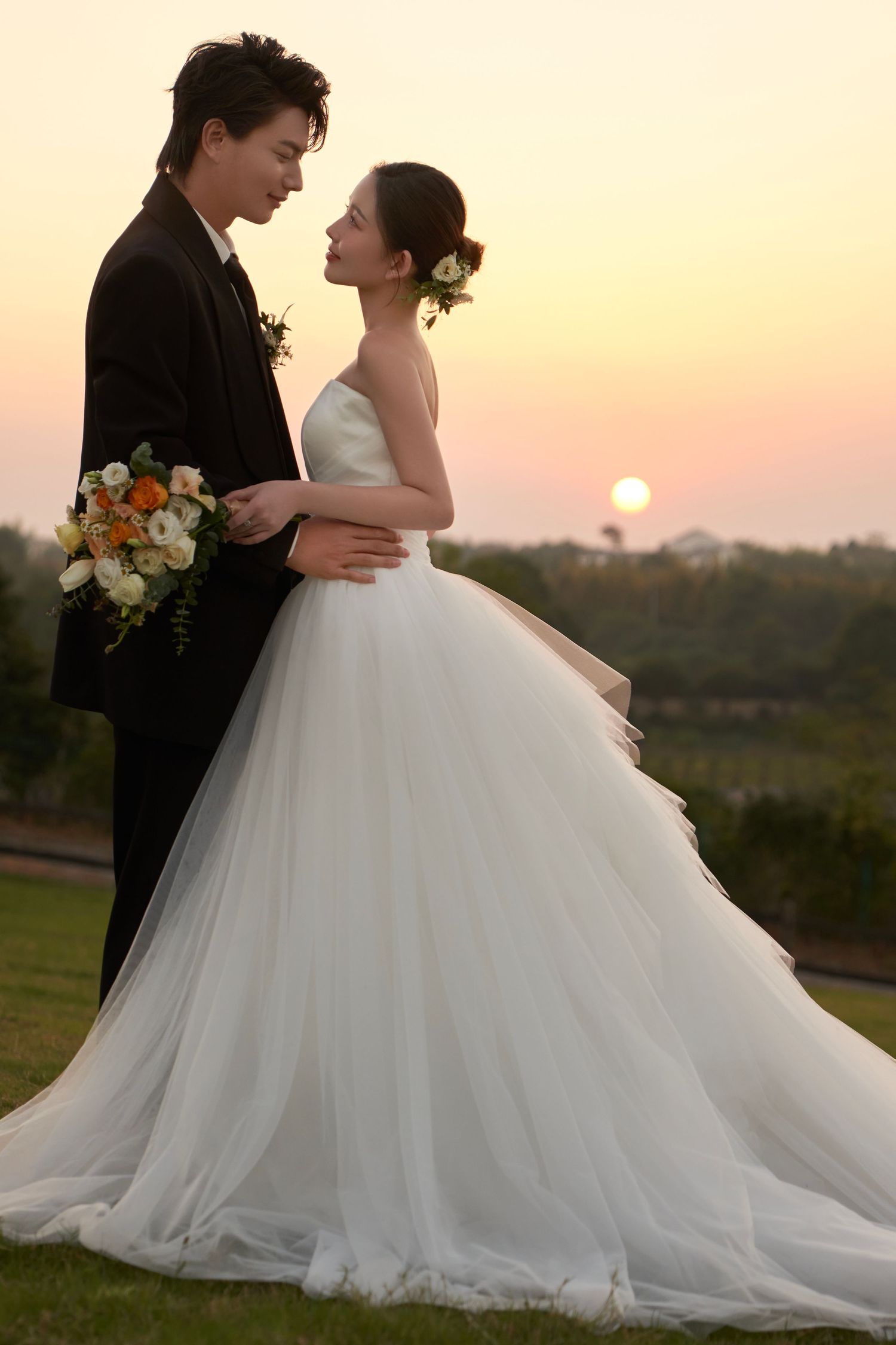 A bride in a white ball gown and groom in a black suit embrace romantically at sunset.