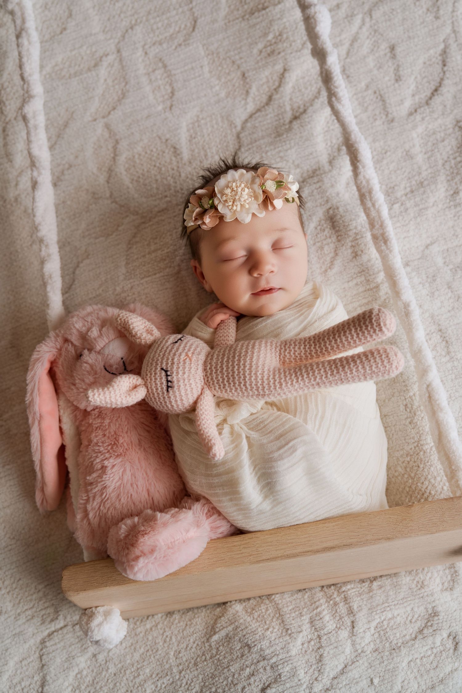 Sleeping newborn baby girl in white swaddle with floral headband, resting beside pink stuffed bunny toys on cream blanket.