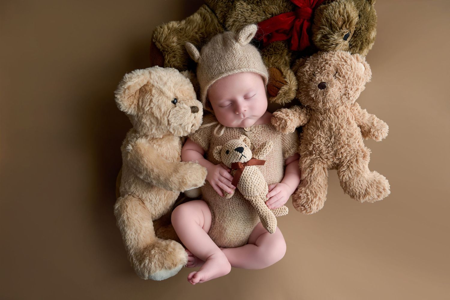 Newborn baby sleeping peacefully between two teddy bears, wearing a knit bear hat, holding a small stuffed bear.