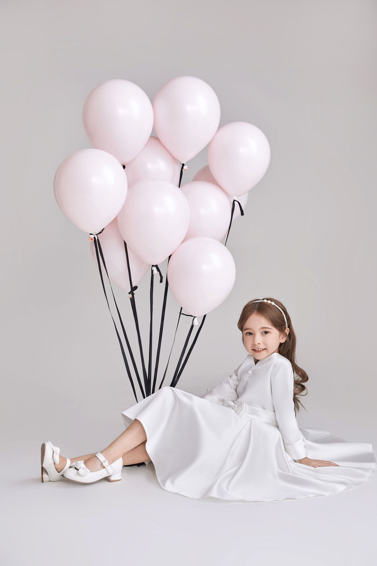 Young girl in white dress sitting on floor with pink balloons in a studio setting.