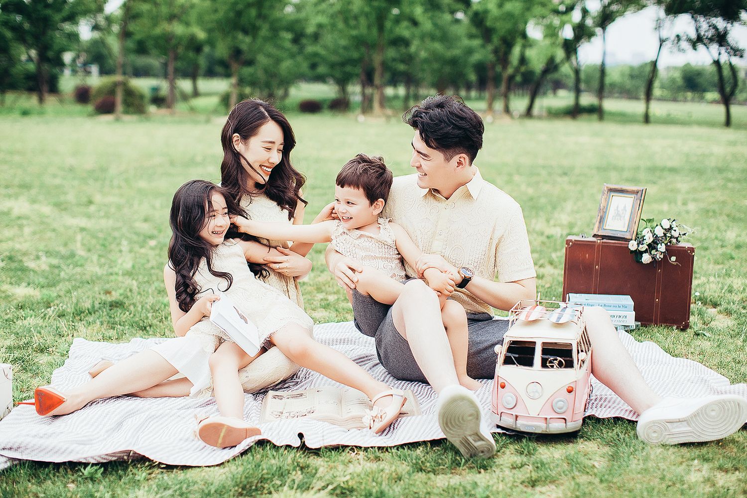 A happy Asian family of four enjoys a picnic on a blanket in a lush green park together.