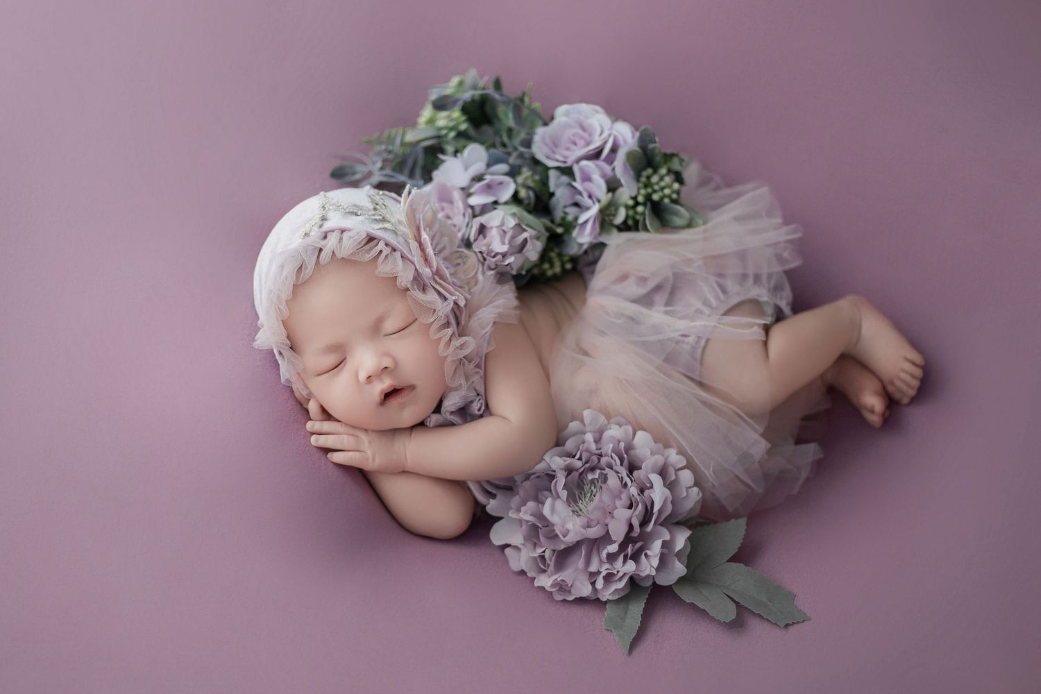 Sleeping newborn baby girl in lavender tutu and floral headband surrounded by purple flowers on pink background.
