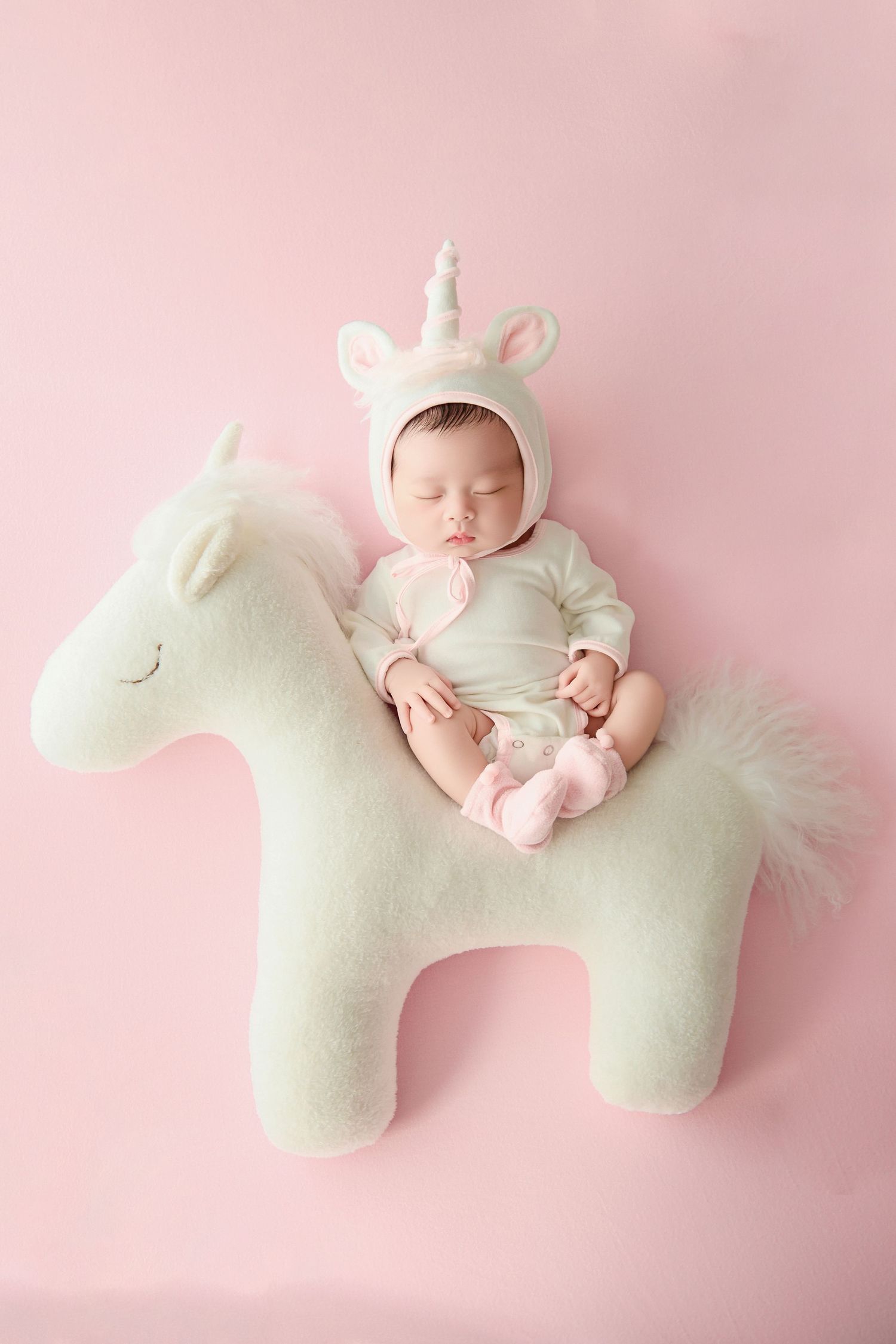 Sleeping newborn baby in unicorn costume lying on white plush unicorn toy against pink background.