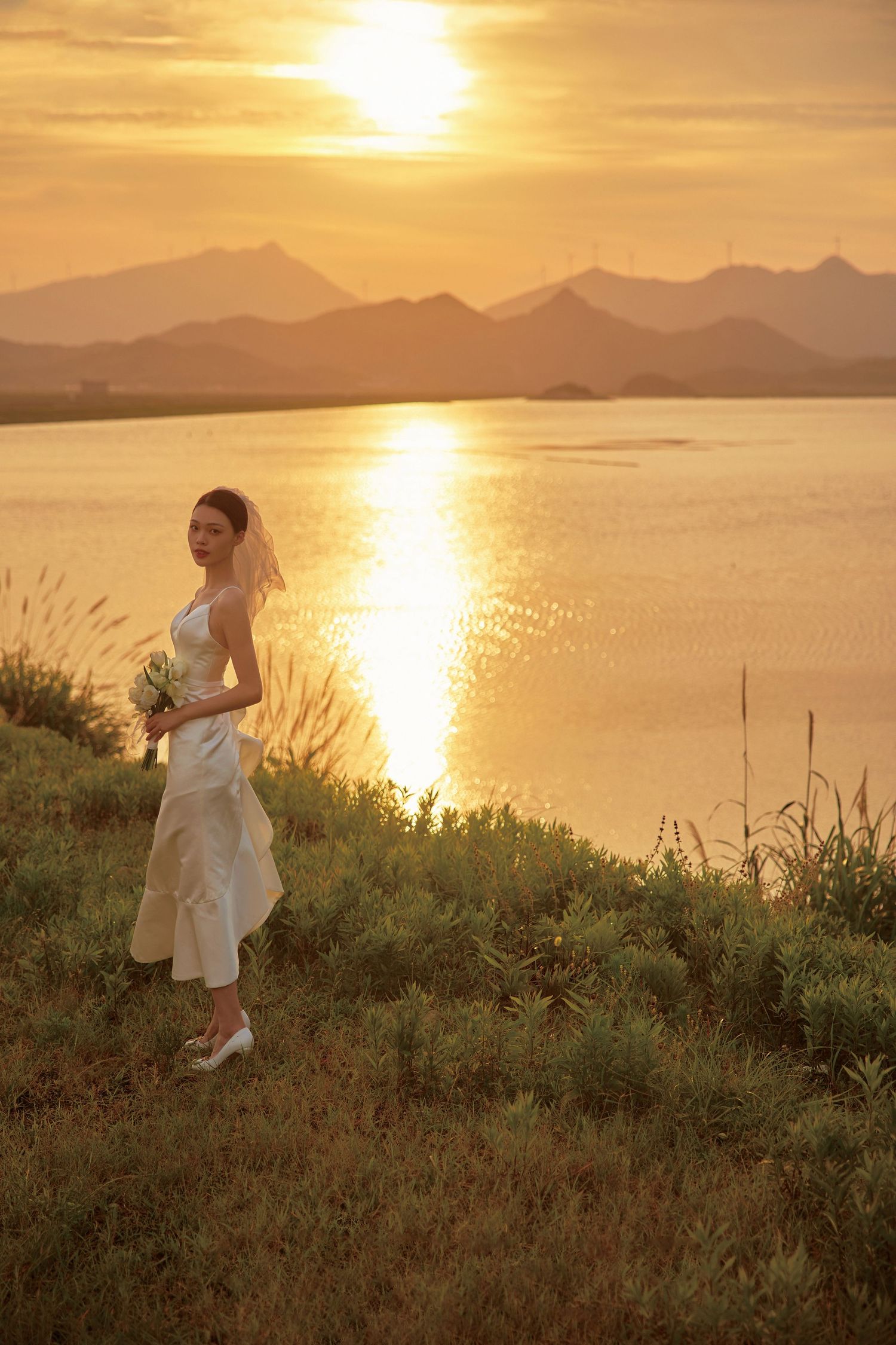 Bride in white gown holding bouquet stands by golden sunset lake with mountains.