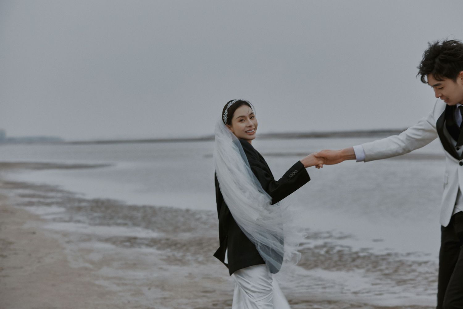 Bride in white veil and black jacket holds groom's hand smiling on overcast misty beach.