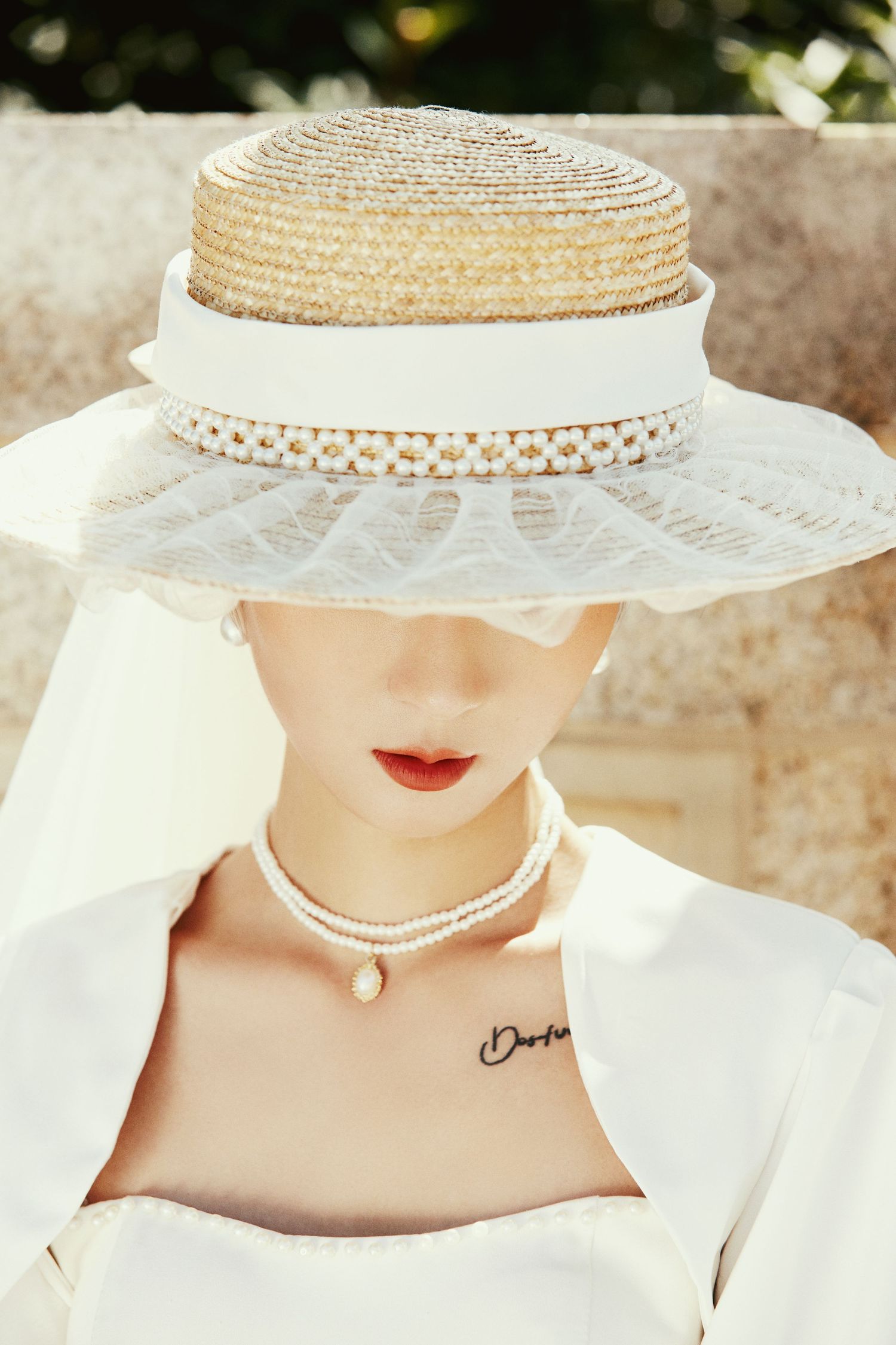 Close-up of bride wearing wide-brim straw hat with veil, pearl necklace, and red lips in outdoor portrait.