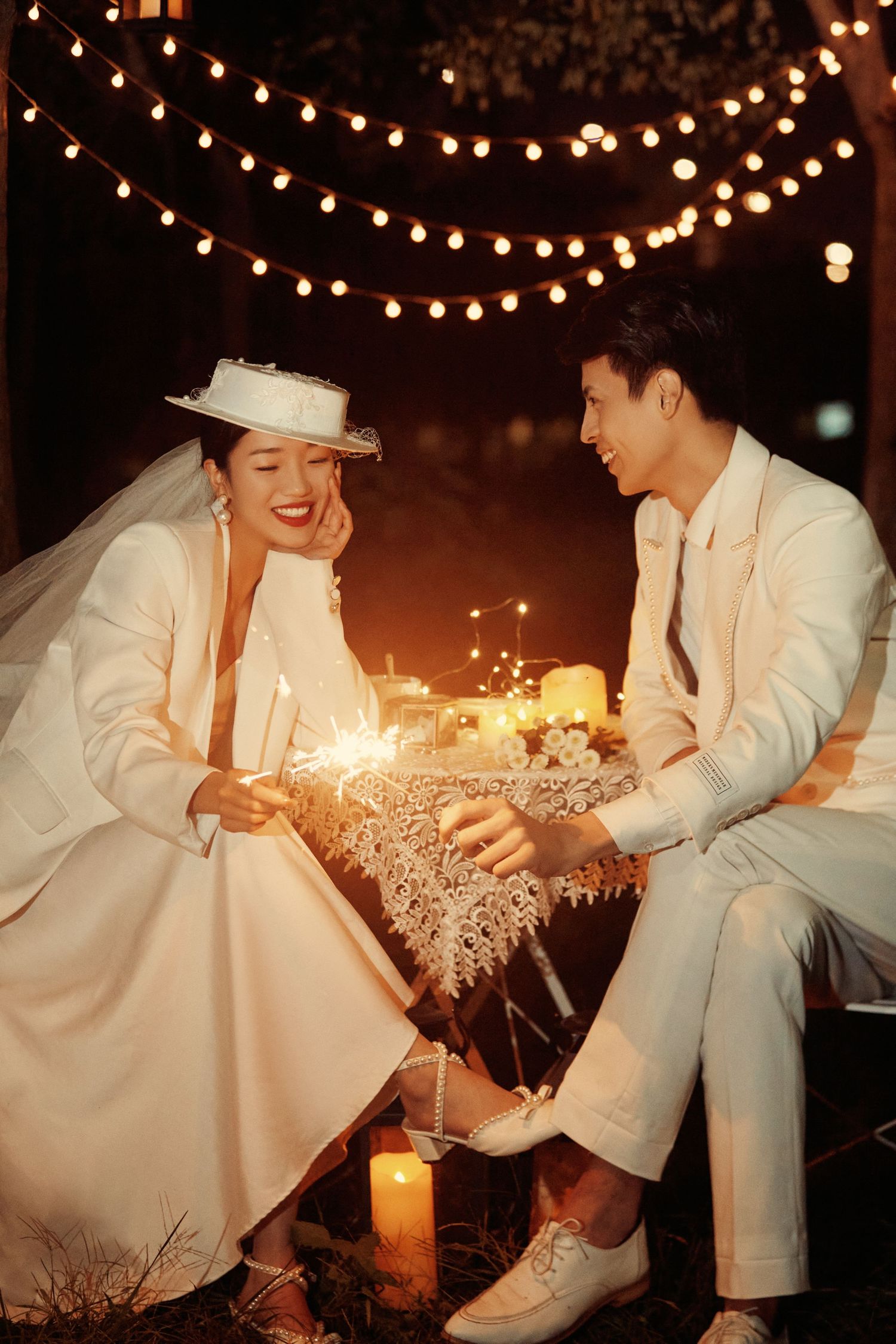 Romantic couple in white wedding outfits holding sparklers by candlelight under string lights at night.
