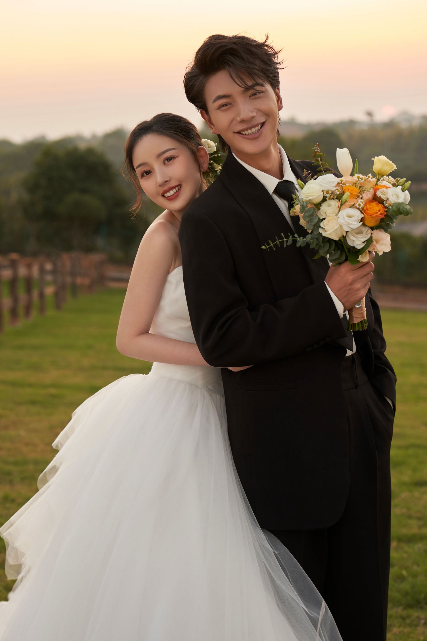 A smiling bride and groom pose back-to-back on a green lawn at sunset, holding a bouquet.