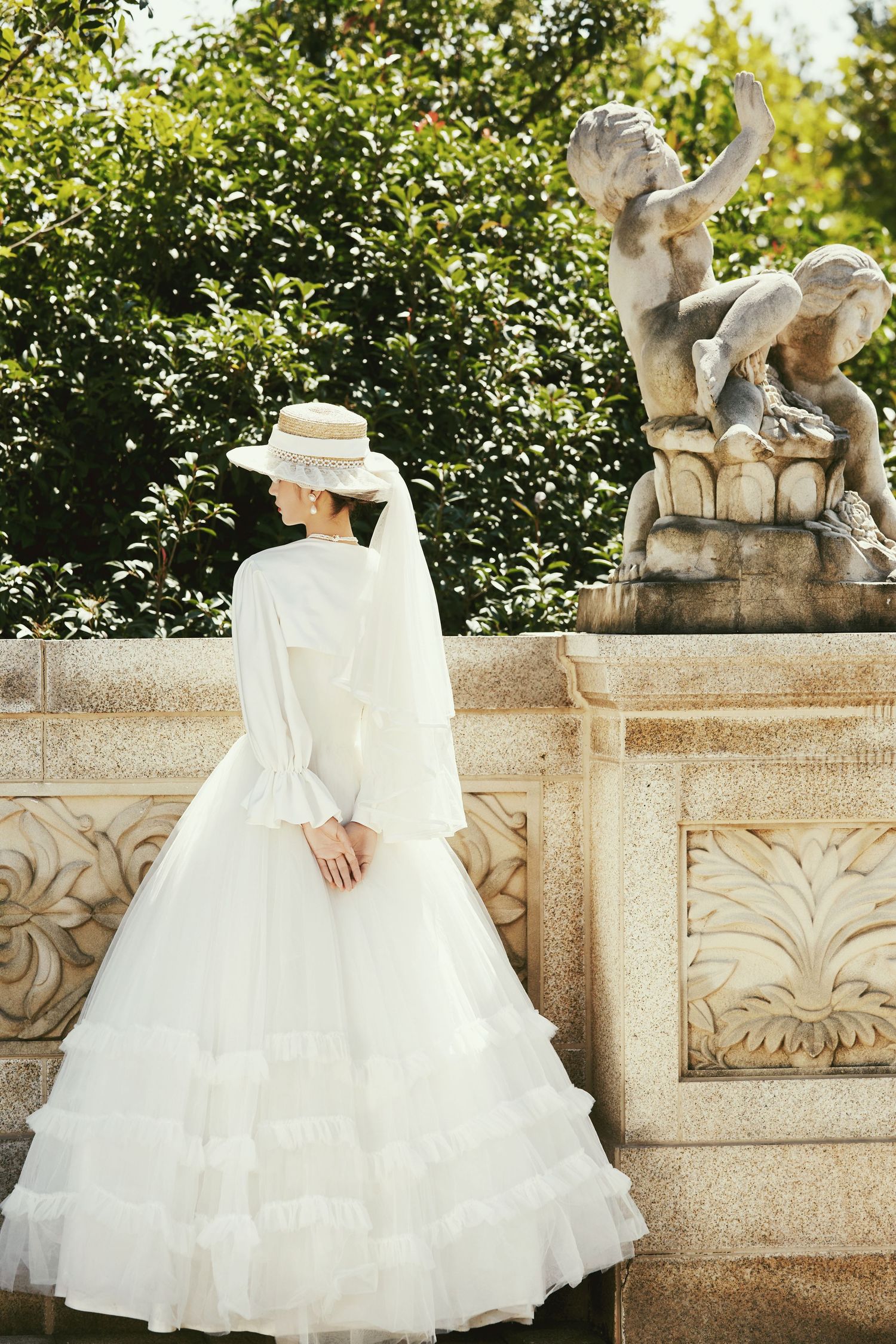 Bride in white ruffled gown and straw hat poses by ornate stone balustrade with classical sculpture in garden.