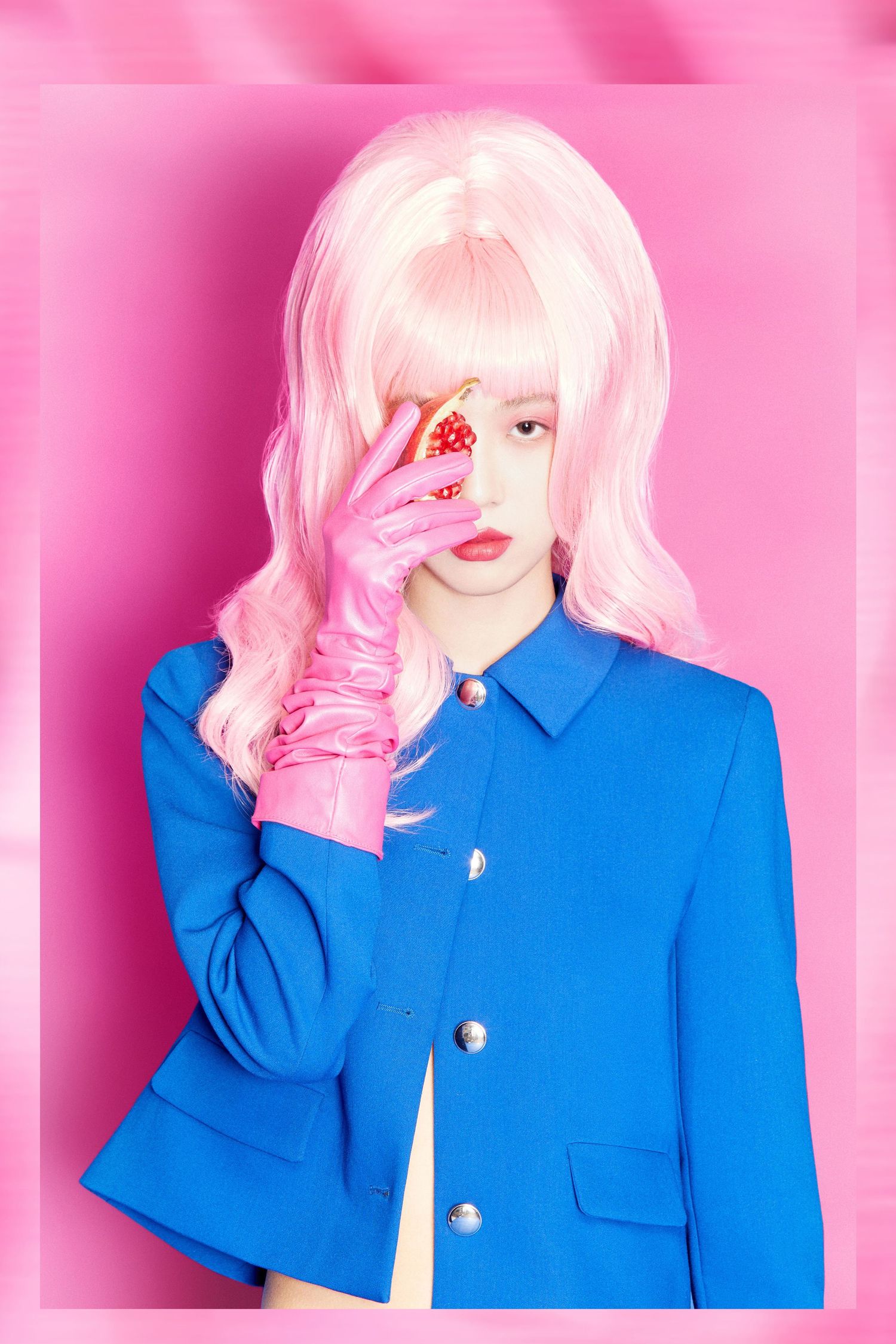 Woman in pink wig and blue blazer holds pomegranate slice over eye, posing against vibrant pink background.
