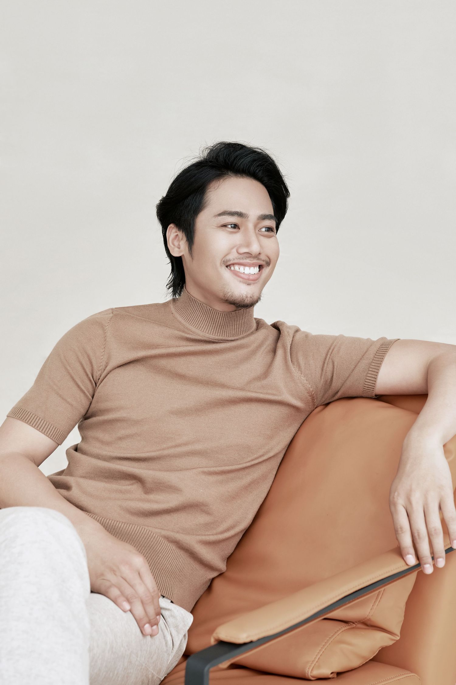 A young Asian man in a beige turtleneck sweater smiling while relaxing in a leather chair.