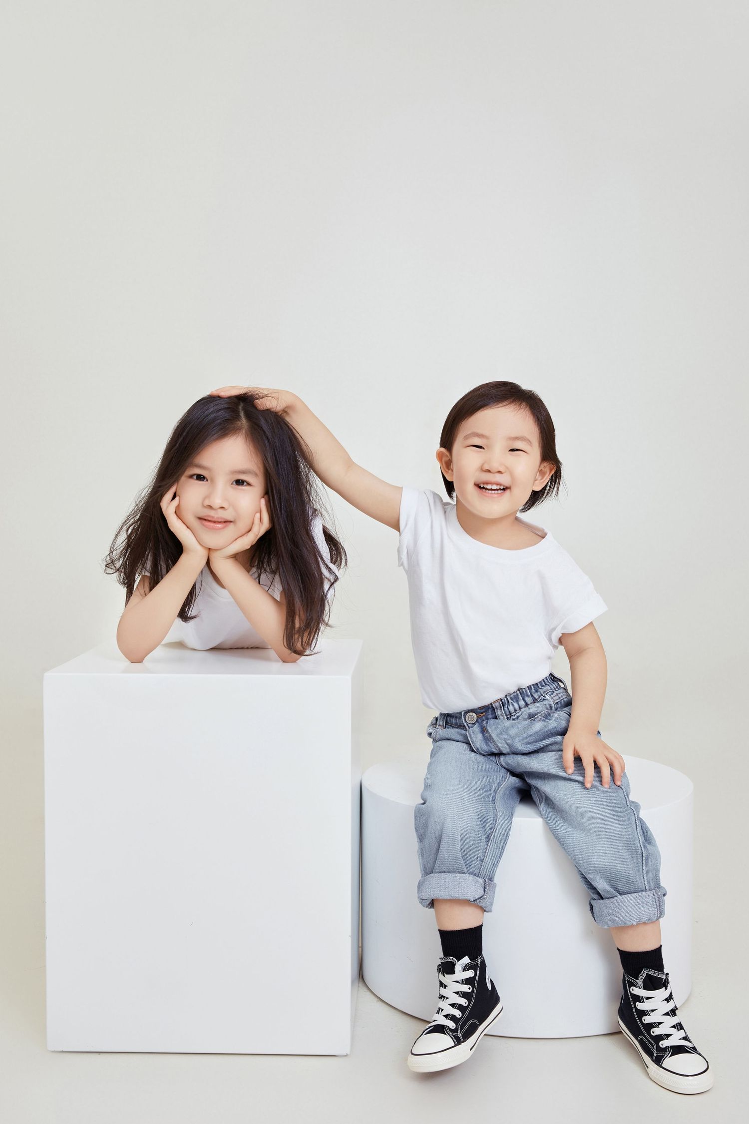 Two adorable Asian siblings in white t-shirts and jeans posing playfully on white cube pedestals in studio.