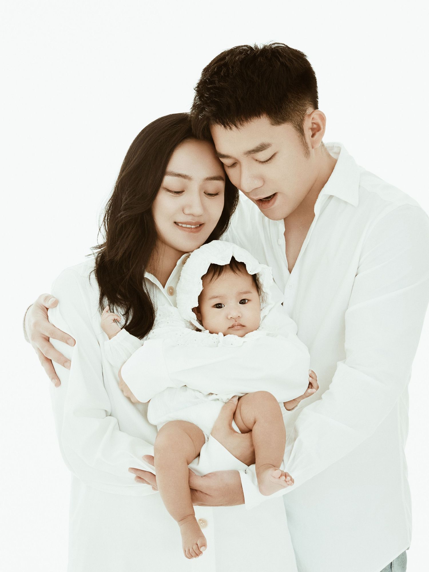 Happy Asian family in matching white outfits pose together holding their newborn baby in a studio.