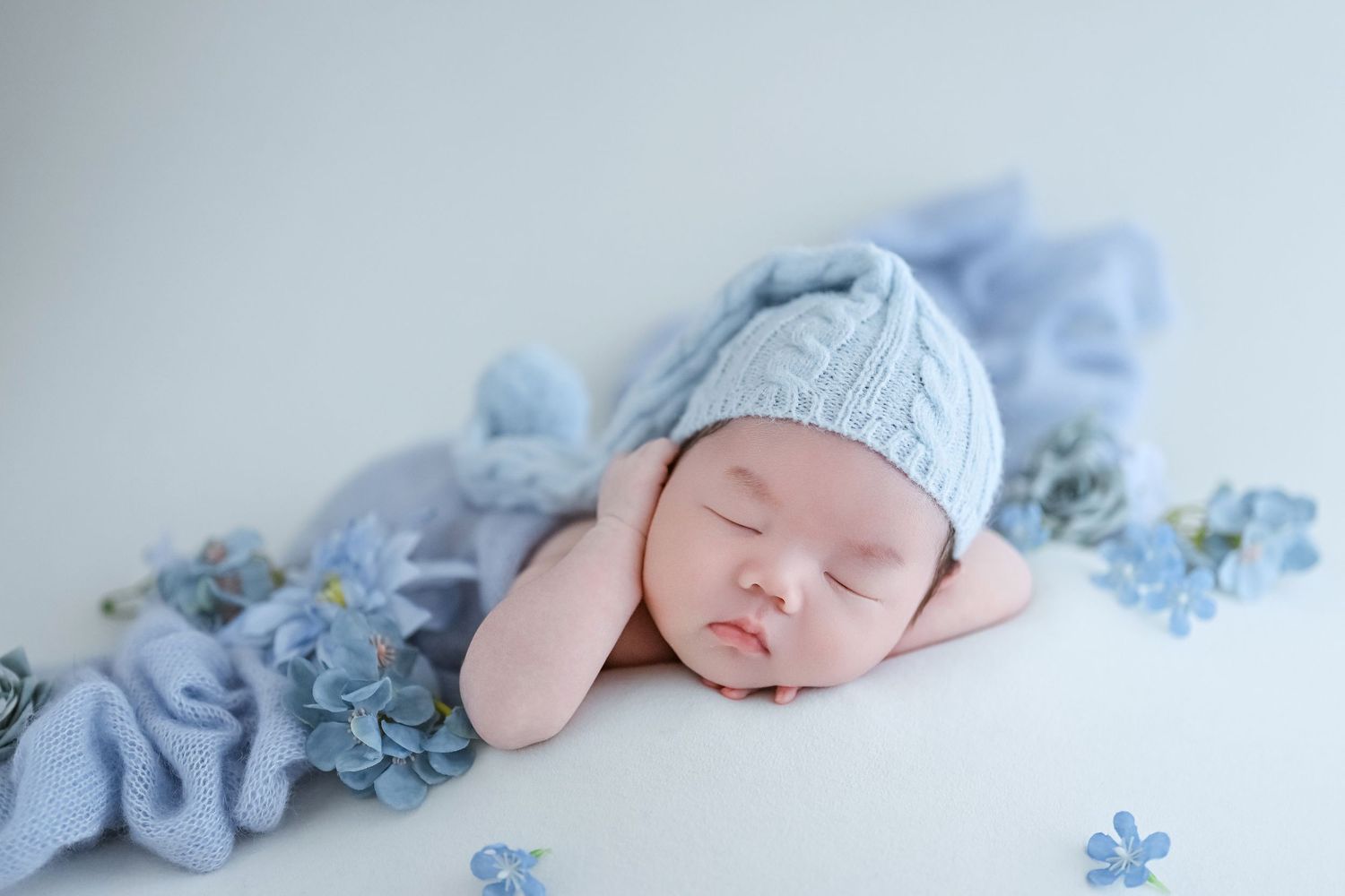 Peaceful newborn baby in blue knit hat sleeping on blue wrap surrounded by delicate blue flowers.