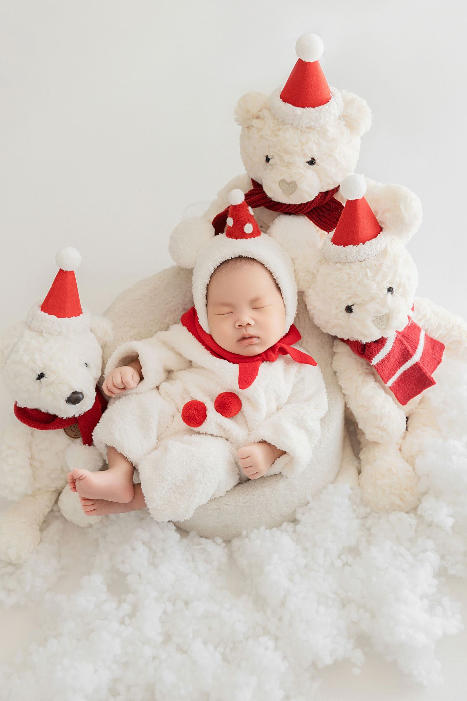 Sleeping newborn baby in white fluffy Santa suit nestled between white Christmas teddy bears on snowy backdrop.