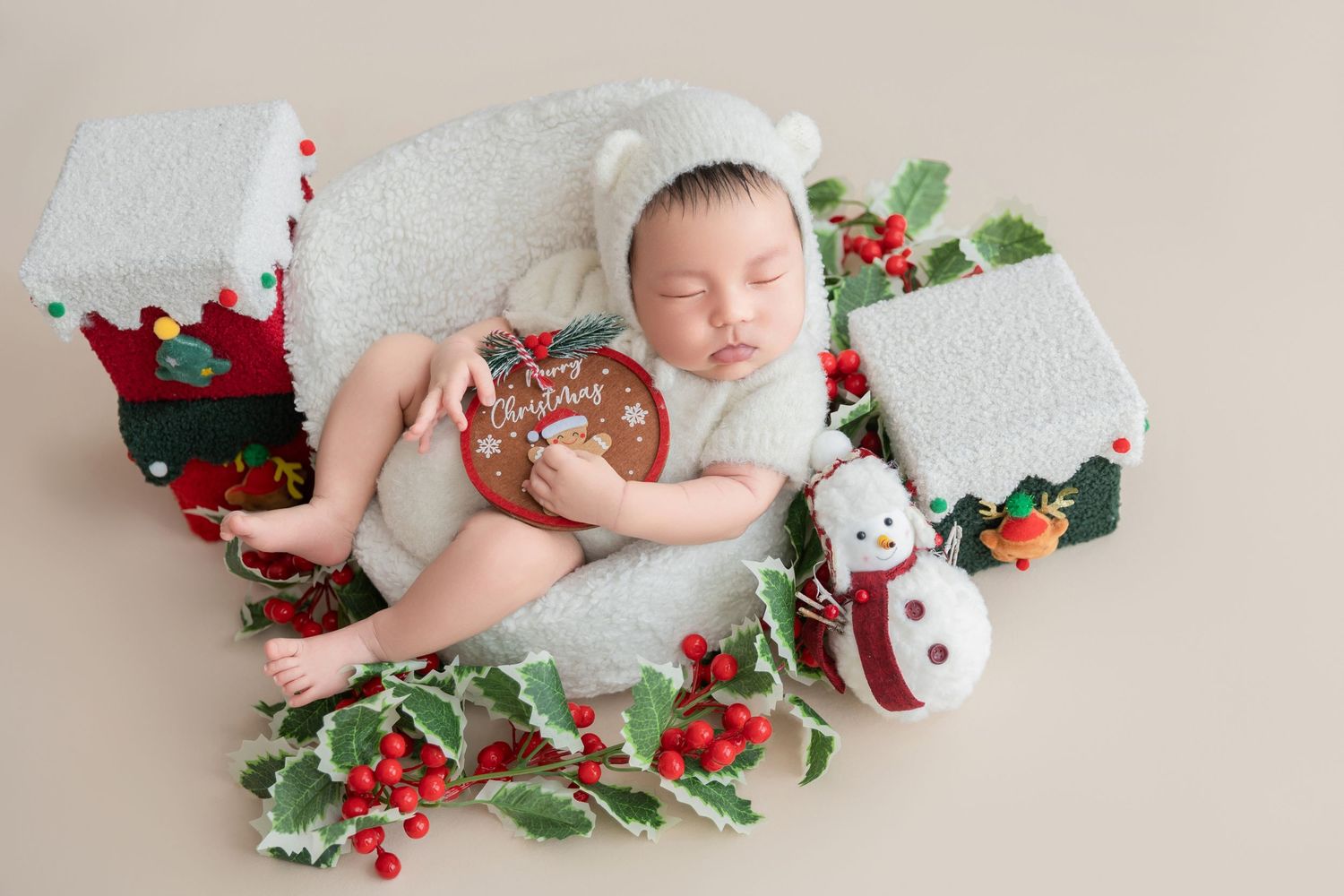 Sleeping newborn in white bear bonnet nestled in holiday greenery with snowman, miniature houses, and red berry decorations.