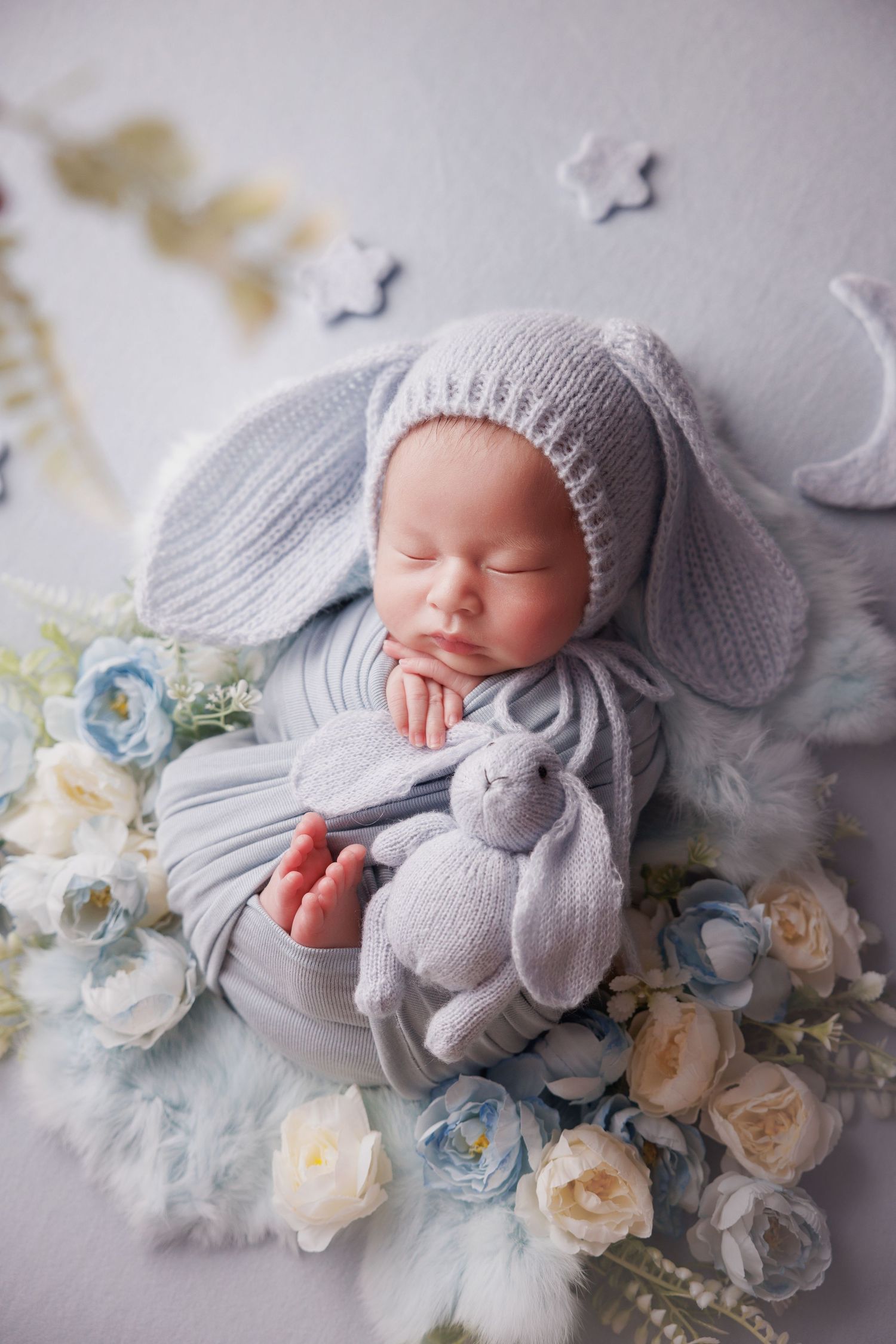 Newborn baby in gray bunny hat cuddling knit rabbit toy surrounded by blue and white roses in cozy bowl.
